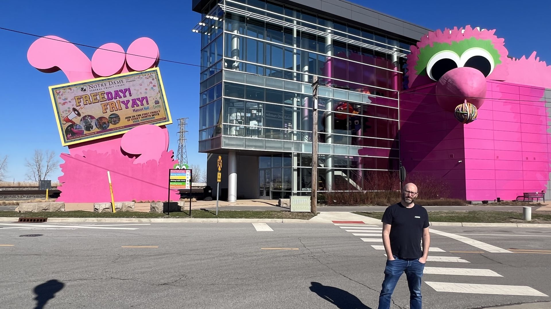 Photo of a man standing in front of a museum with a huge pink cartoon face on it. 