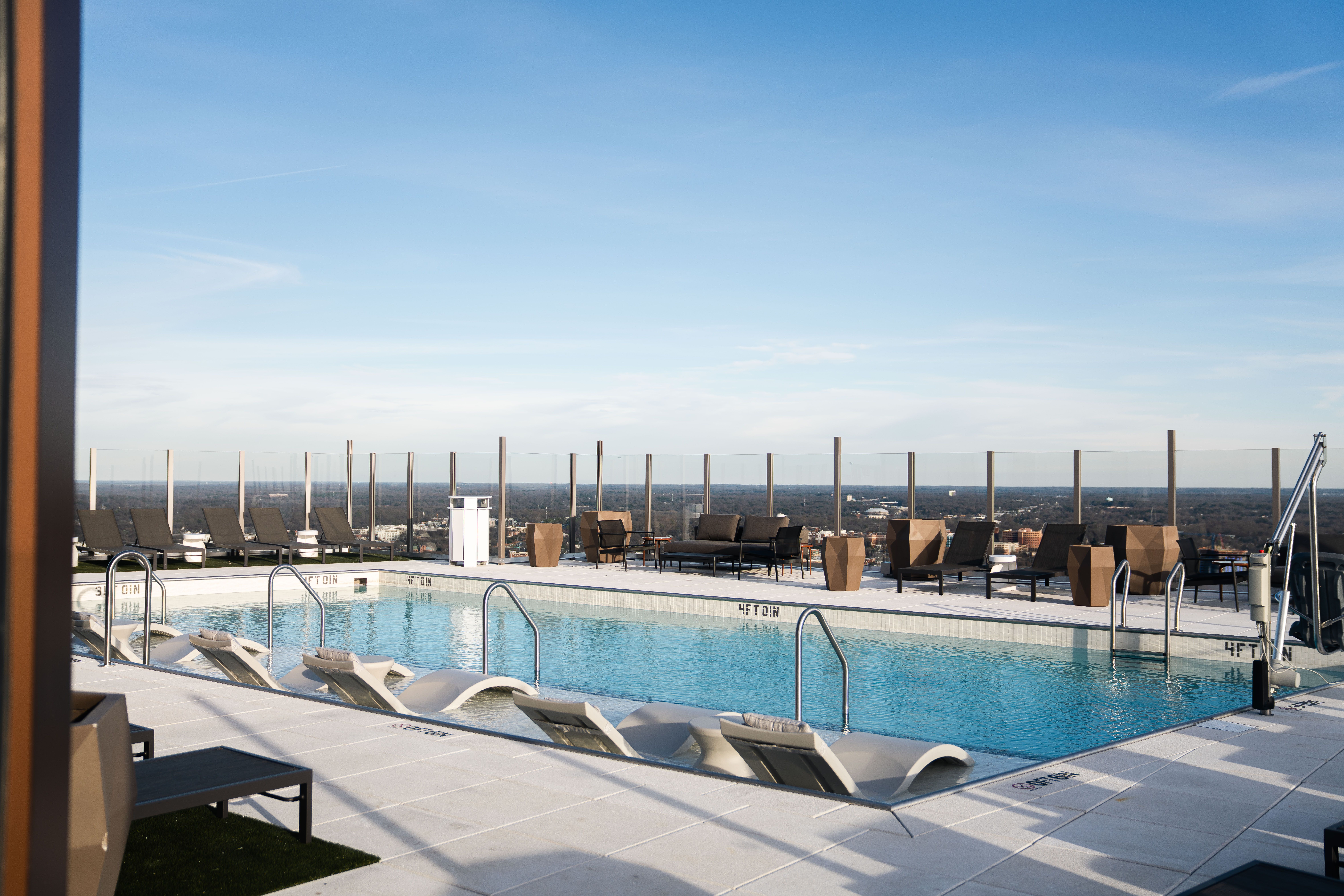 Rooftop swimming pool with curved white lounge chairs partially submerged in water, brown cushioned chairs, glass safety barriers, and clear blue sky over a cityscape in the background.