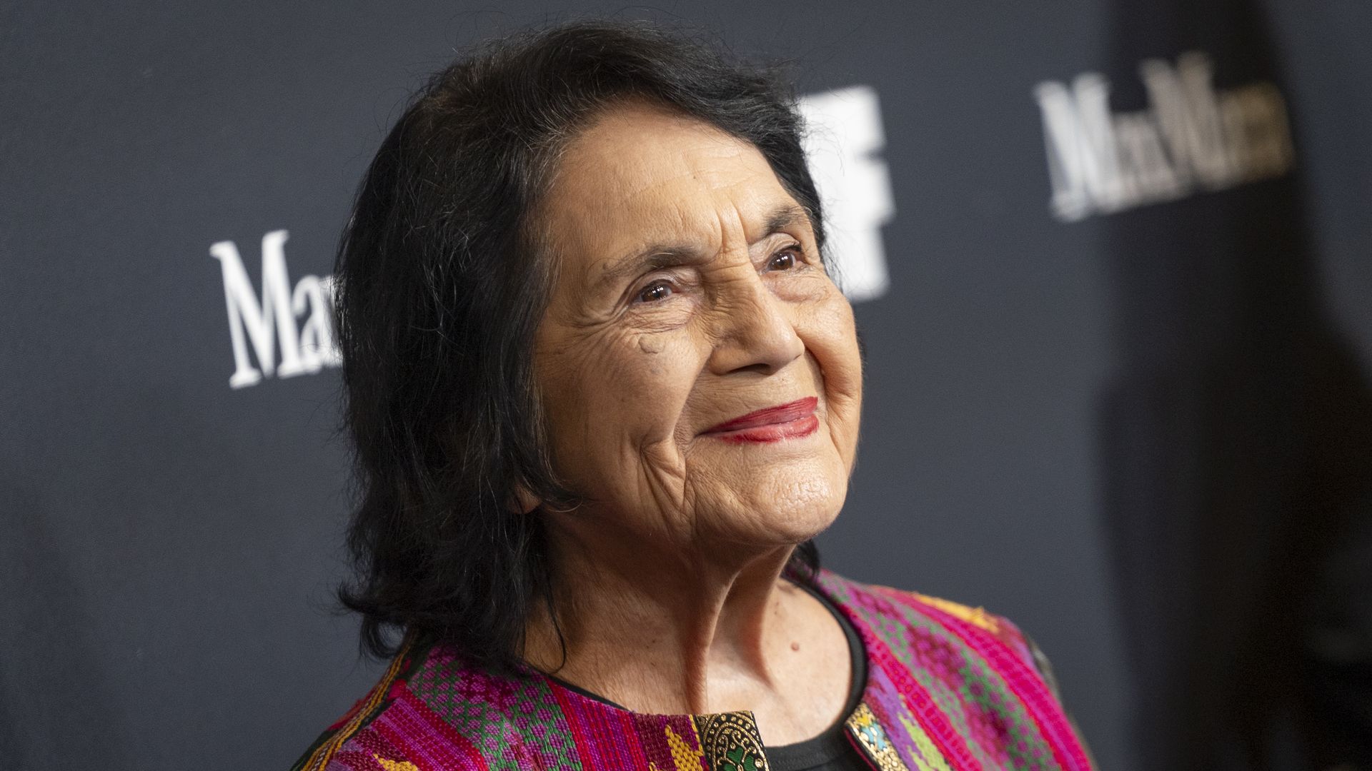 Close-up of an older woman with short black hair and red lipstick, wearing a colorful pink, green, and gold patterned jacket, smiling softly against a dark backdrop.