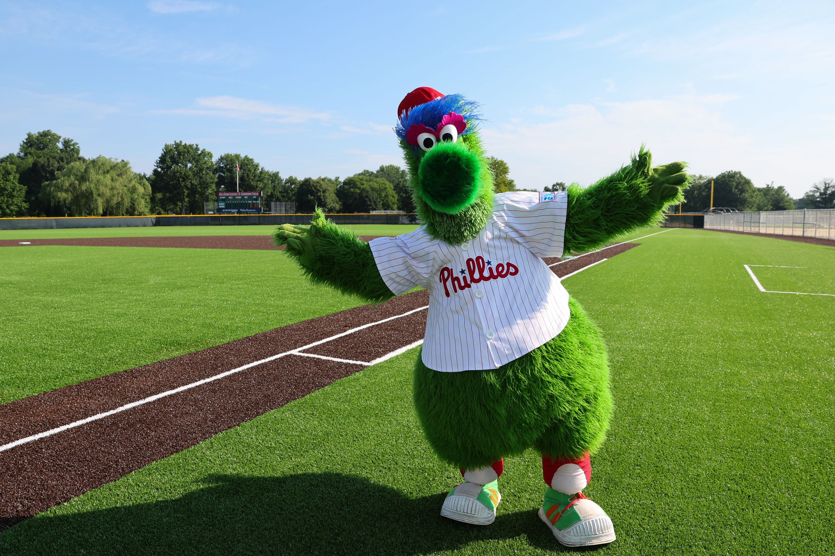 Bright green baseball mascot in a Phillies jersey with red lettering stretches arms on a sunny baseball field; blue and red hair, large eyes, green snout, with infield dirt and distant scoreboard.