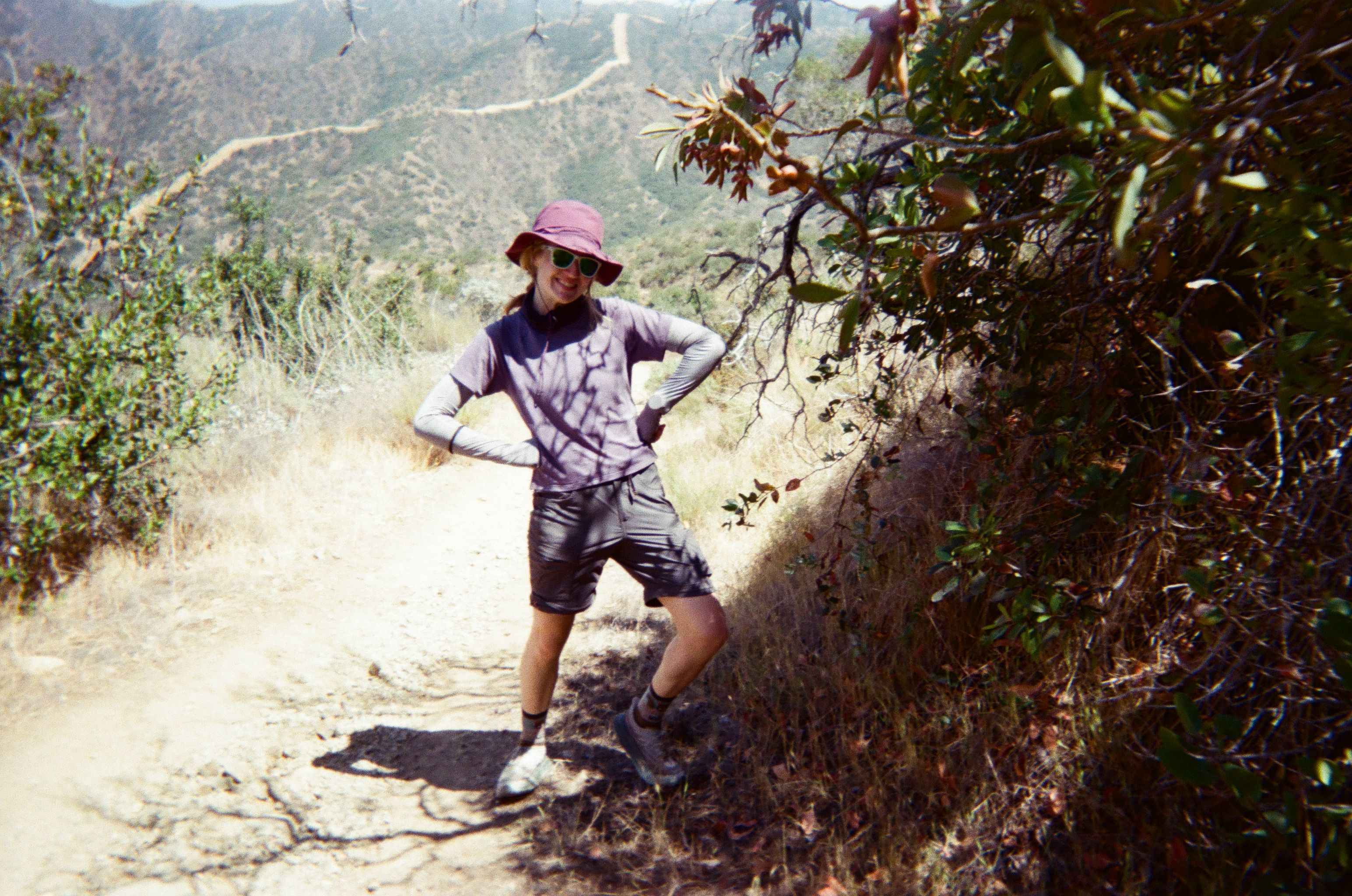 Hiker wearing a pink bucket hat and sunglasses stands with hands on hips on a dusty, sunlit trail in a dry, hilly landscape; a leafy bush frames the right and distant hills wind in the background.