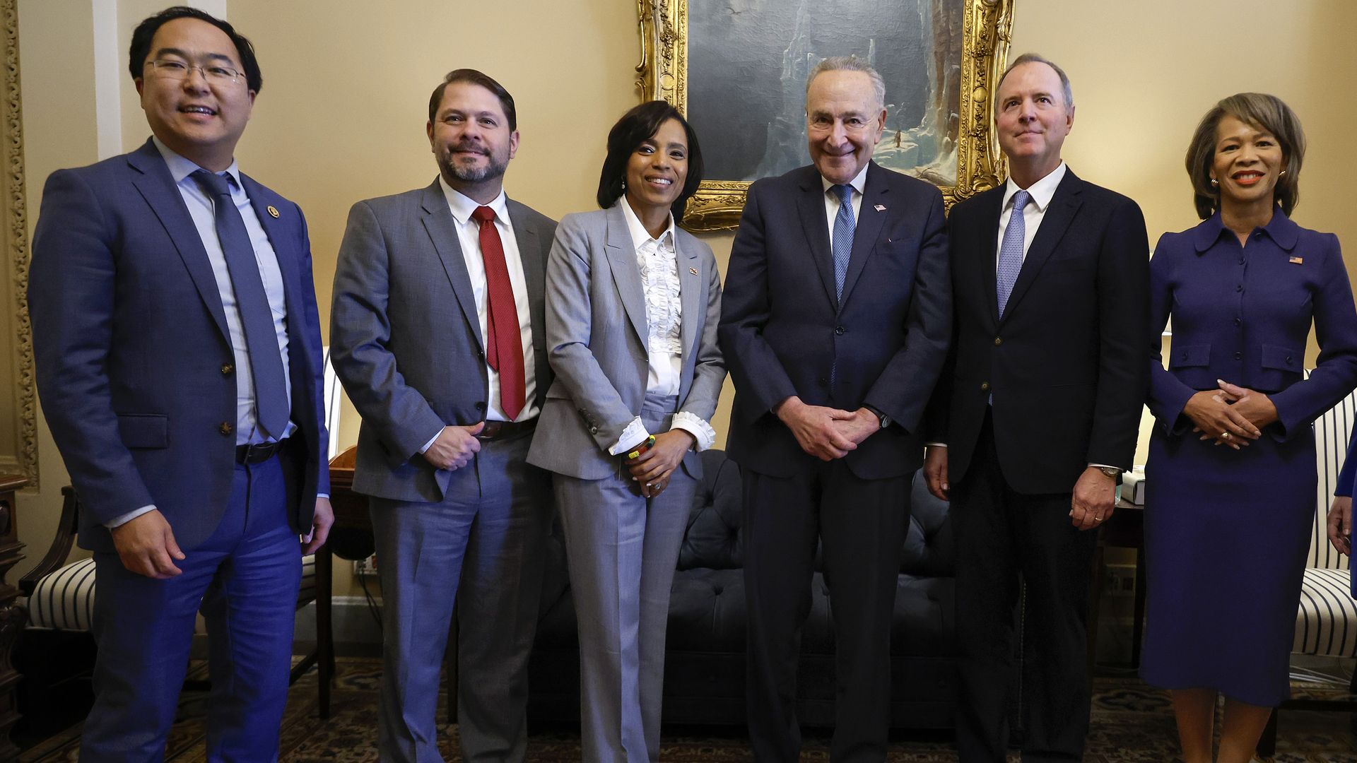 Six people in professional attire standing together, four men and two women, in an official looking room, in front of a gilt-framed painting. 