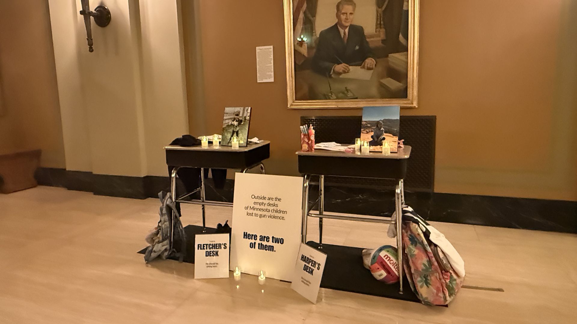 Two school desks displayed with candles, photos, backpacks, and signs in a room with beige walls and a portrait. Signs highlight Minnesota children lost to gun violence.