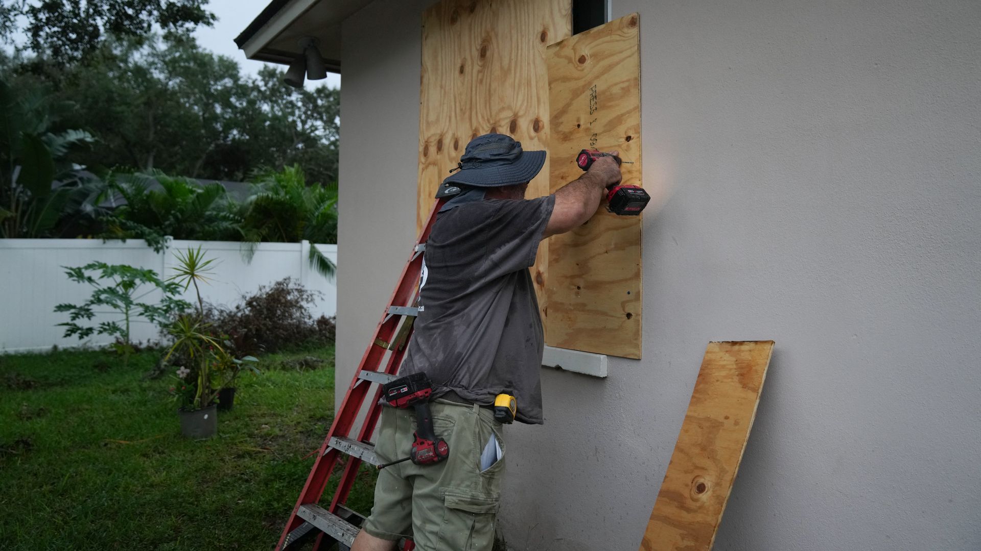 Man in shorts and hat using a drill to secure plywood over a window on a beige house, with a red ladder and greenery in the background during overcast weather.