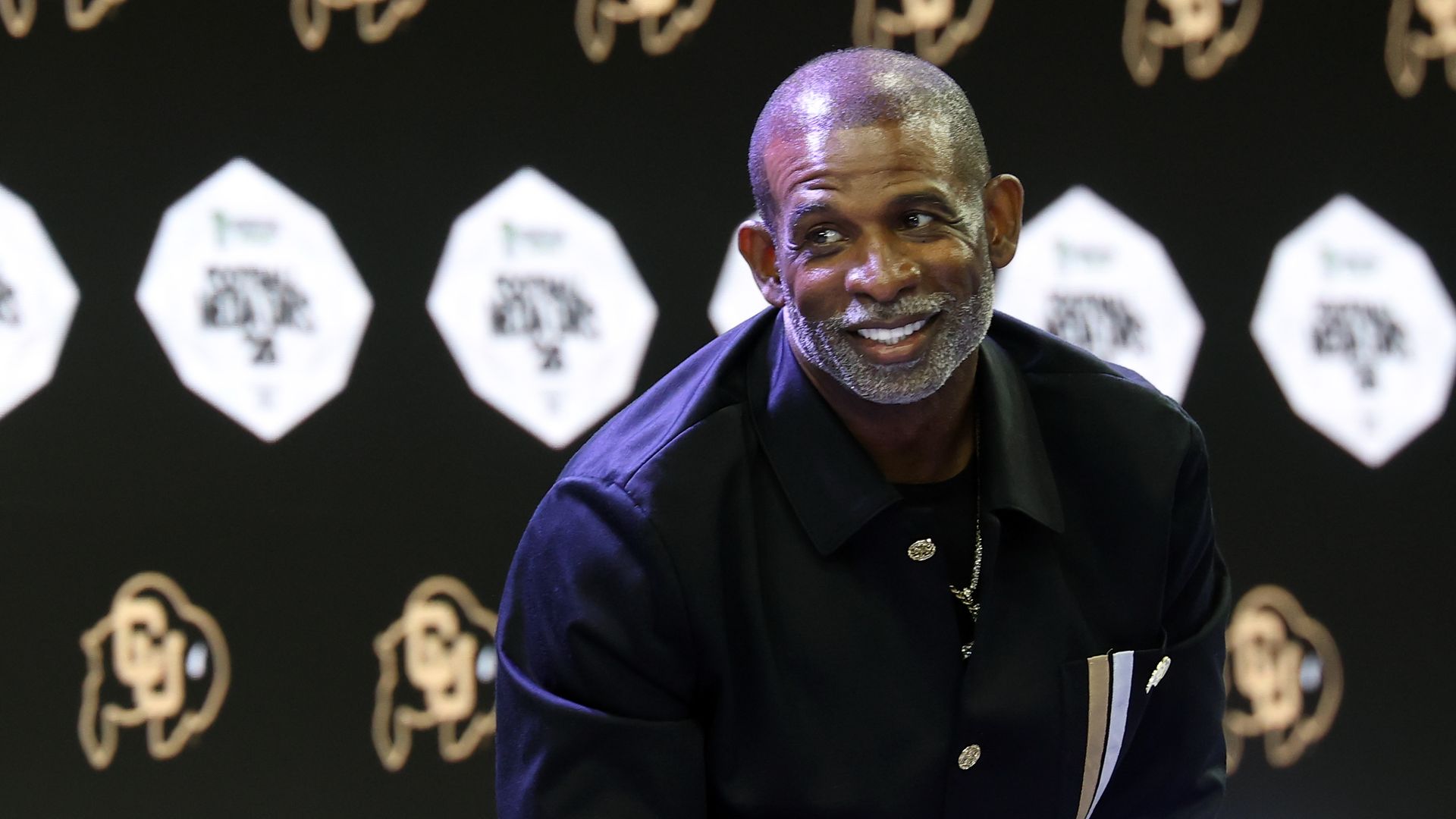 Smiling middle-aged man with a gray beard in a dark suit, standing in front of a black backdrop with gold emblems and white diamond banners at a press event.