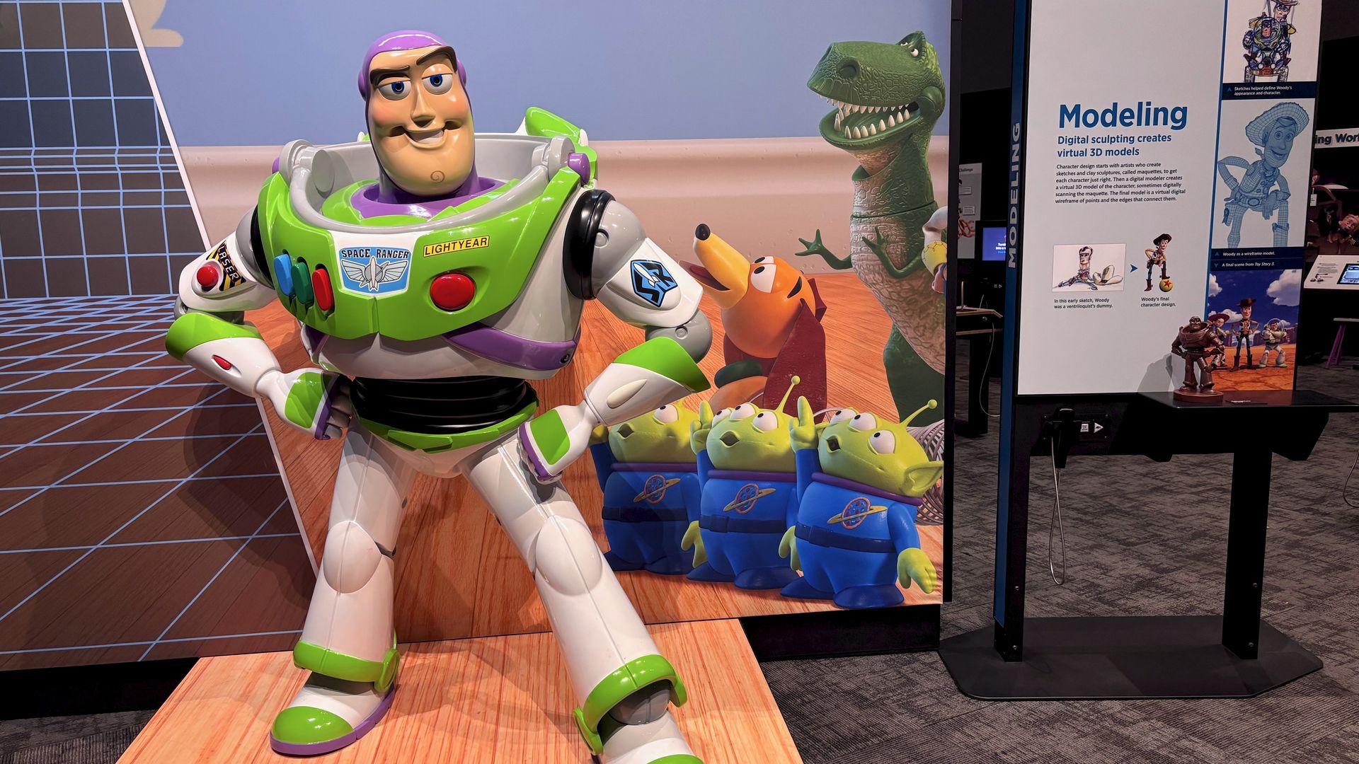 Life-size Buzz Lightyear figure in white and green space suit stands on a wooden platform in front of Toy Story character mural. Nearby is a display board titled "Modeling" explaining digital sculpting.