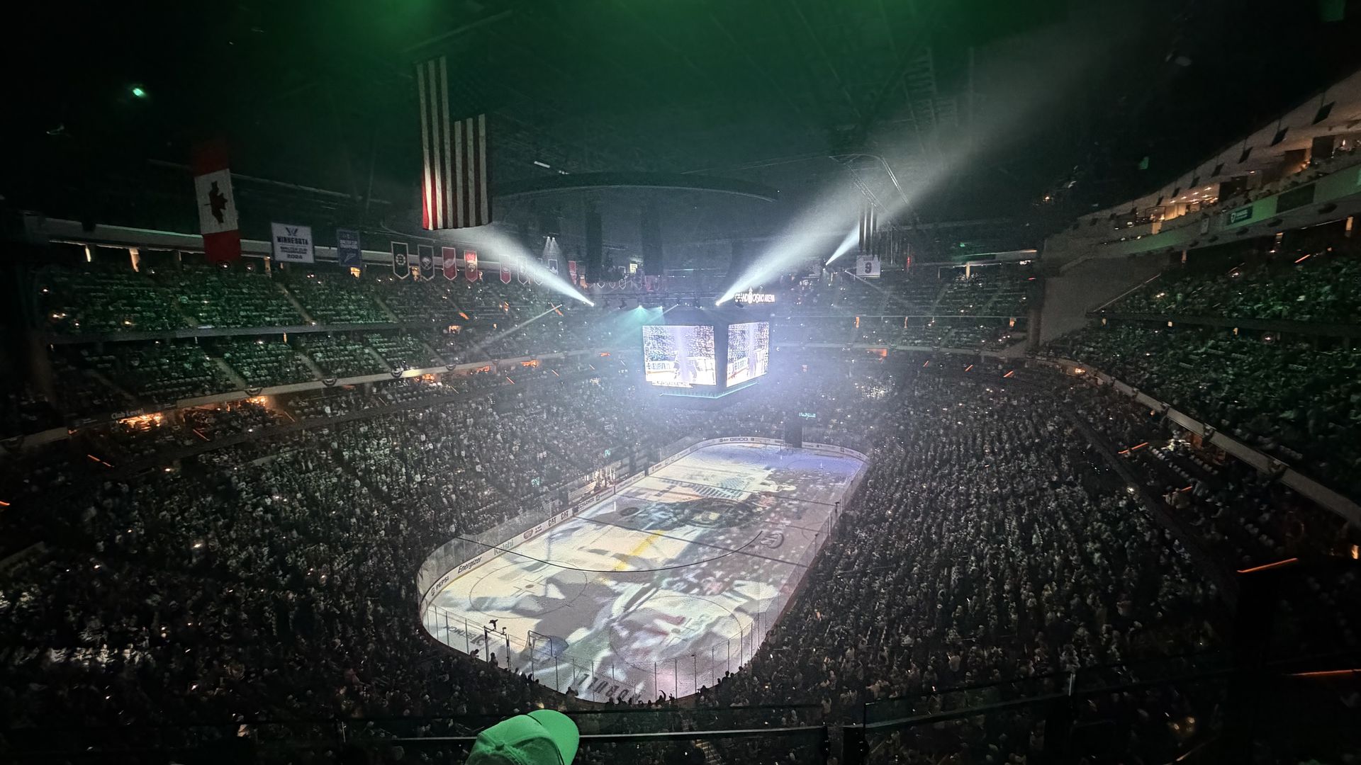 Packed indoor hockey arena full of fans. Center ice with large screens and bright spotlights; banners and flags, including the U.S. and Canadian flags, hang from the upper levels.