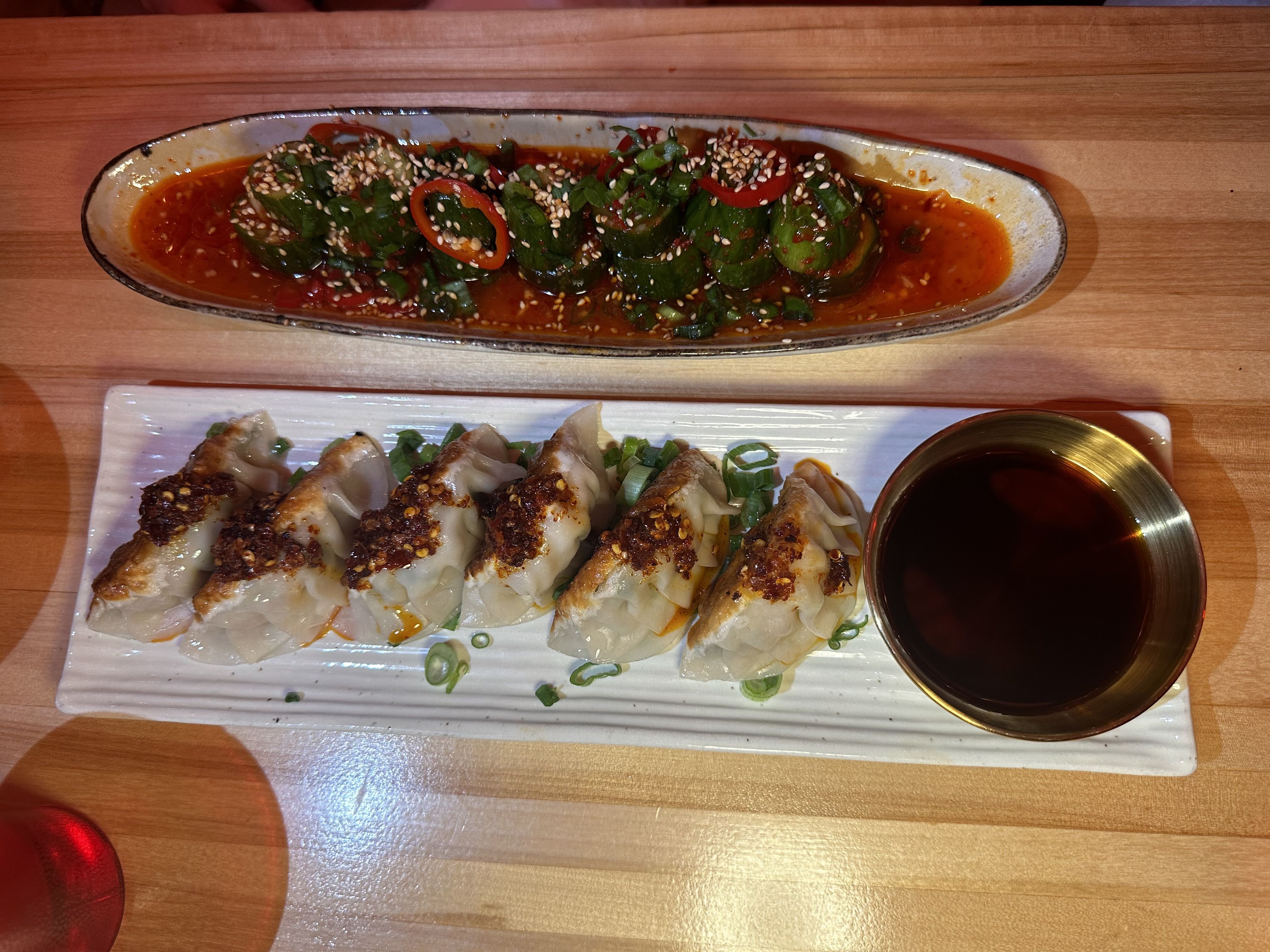 Long white plate with pan-fried dumplings in red chili oil, sesame seeds, green onions, and red chili slices. A soy sauce cup sits on a wooden table.
