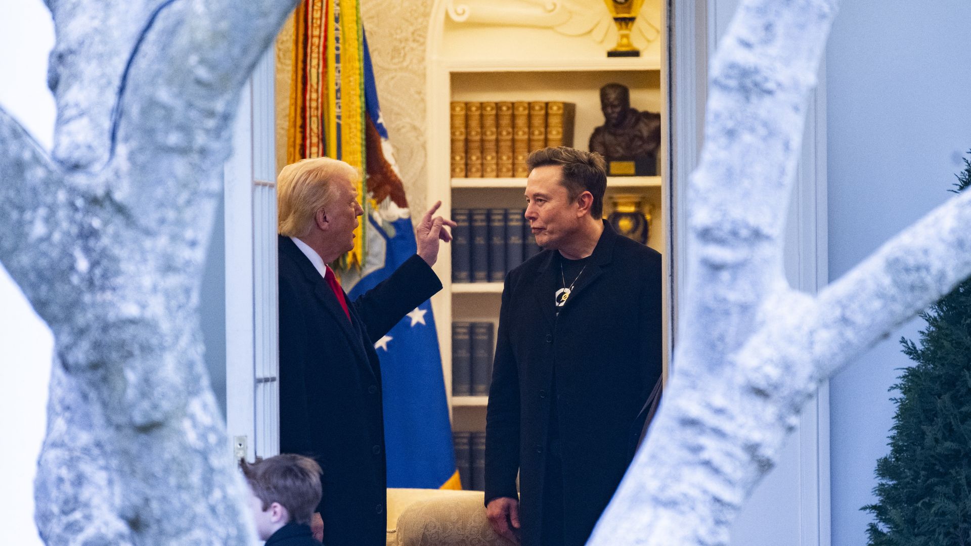 President Trump raises his finger while talking with Elon Musk in the Oval Office in a photo framed by a tree outside the office.