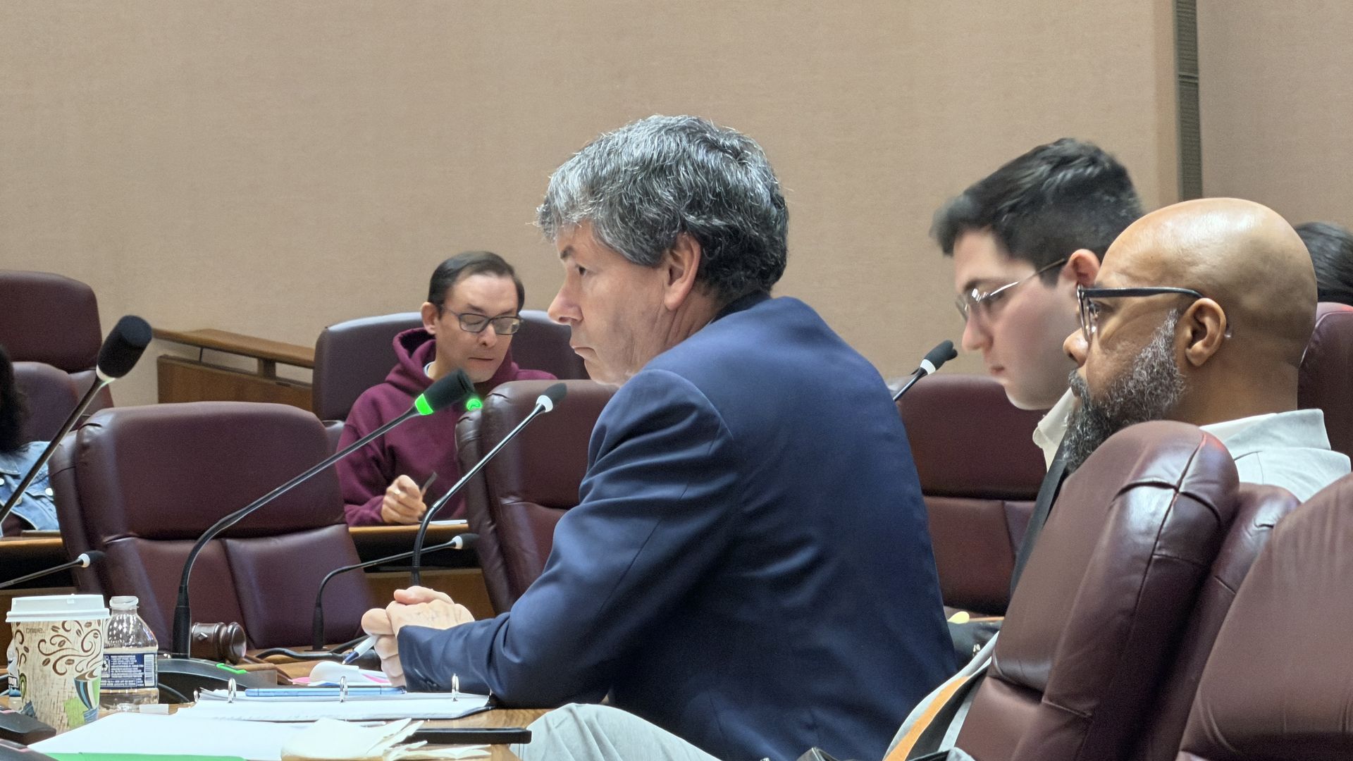 Four men seated in a meeting room with brown leather chairs and wooden table; one man in a navy blazer speaking into a microphone, others listening; coffee cup, water bottle, and papers on table.