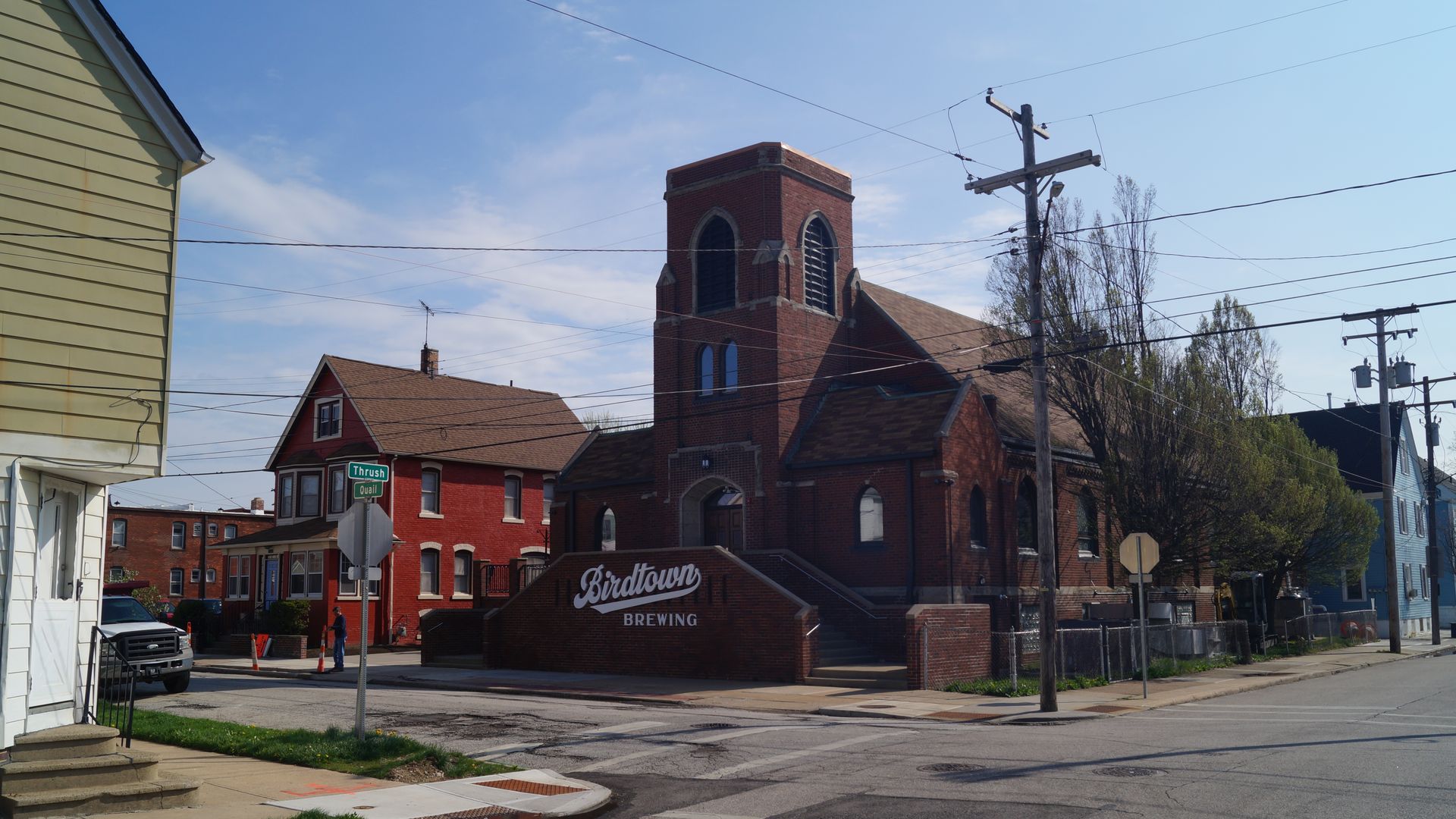Sunny street corner with a brick building labeled "Birdtown Brewing" near red brick houses; a tall brick church-like structure with a tower; power lines cross the blue sky.