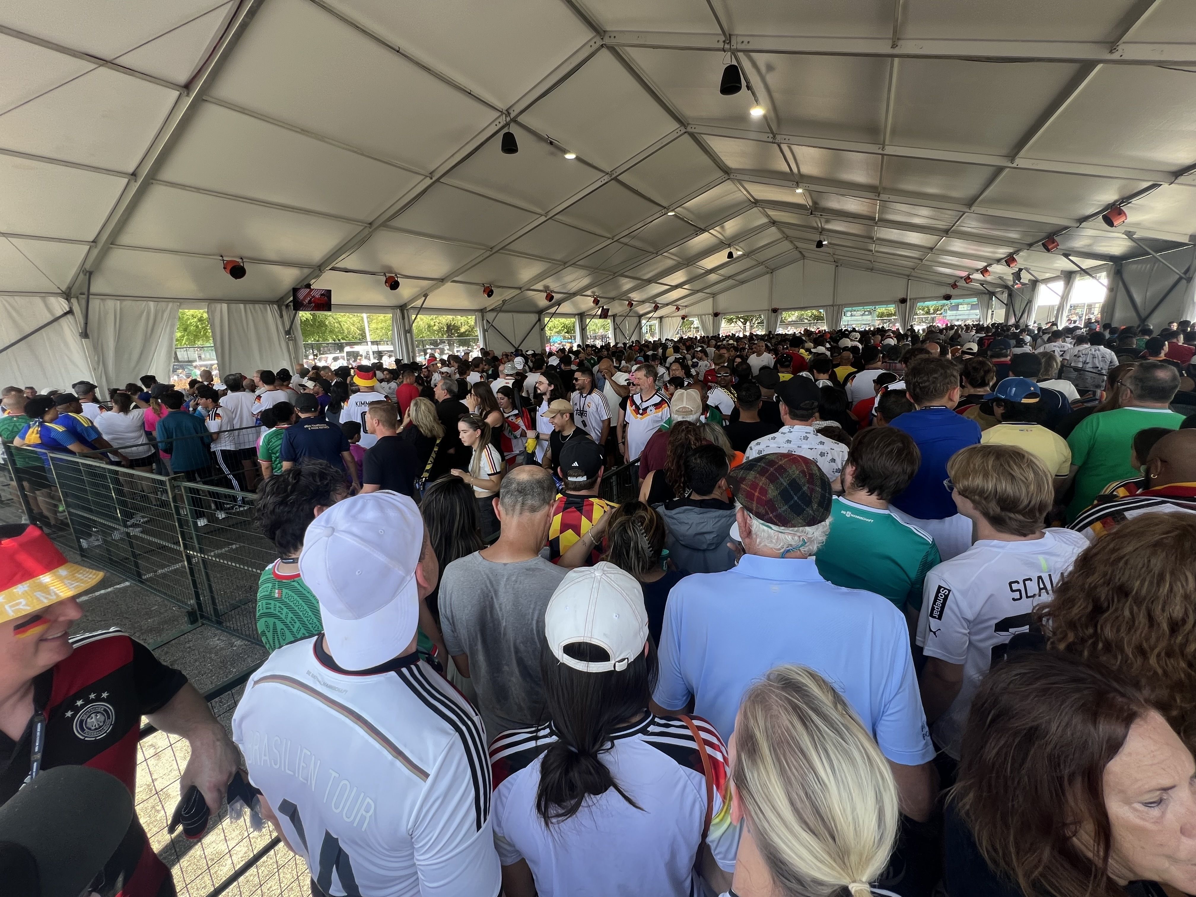 A crowded event tent filled with fans in sports jerseys and caps, standing behind metal barriers; overhead lights hang from the white framework, with small screens along the walls.