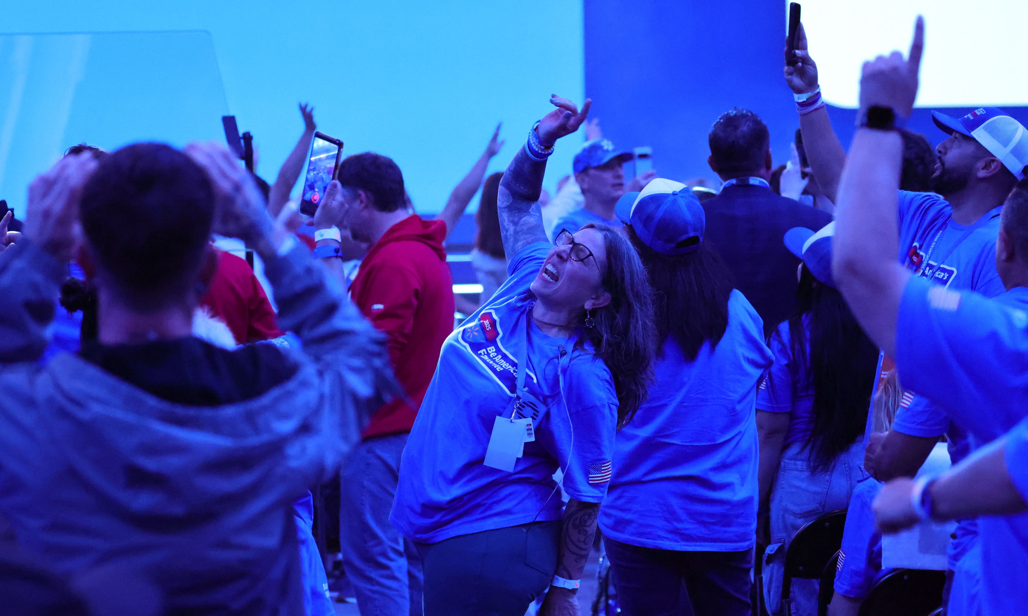 Excited woman in blue Walmart shirt dances joyfully in a crowd, surrounded by people filming with phones under blue lights.