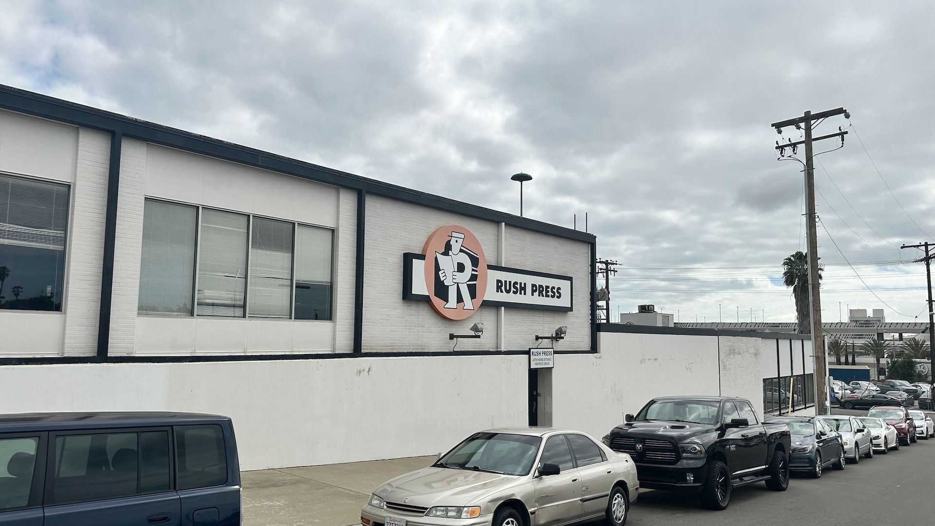 A "Rush Press" building downtown with cars parking in front along the road. 