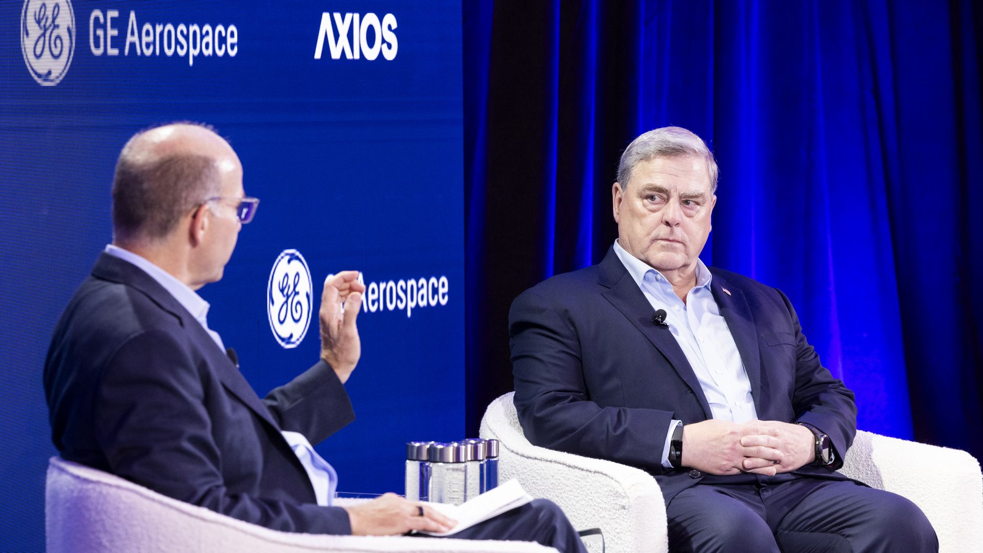 Two men, Mike Allen and Mark Milley, wearing blue suits are seated in white, fuzzy chairs on a stage. Blue dominates the background, as well.