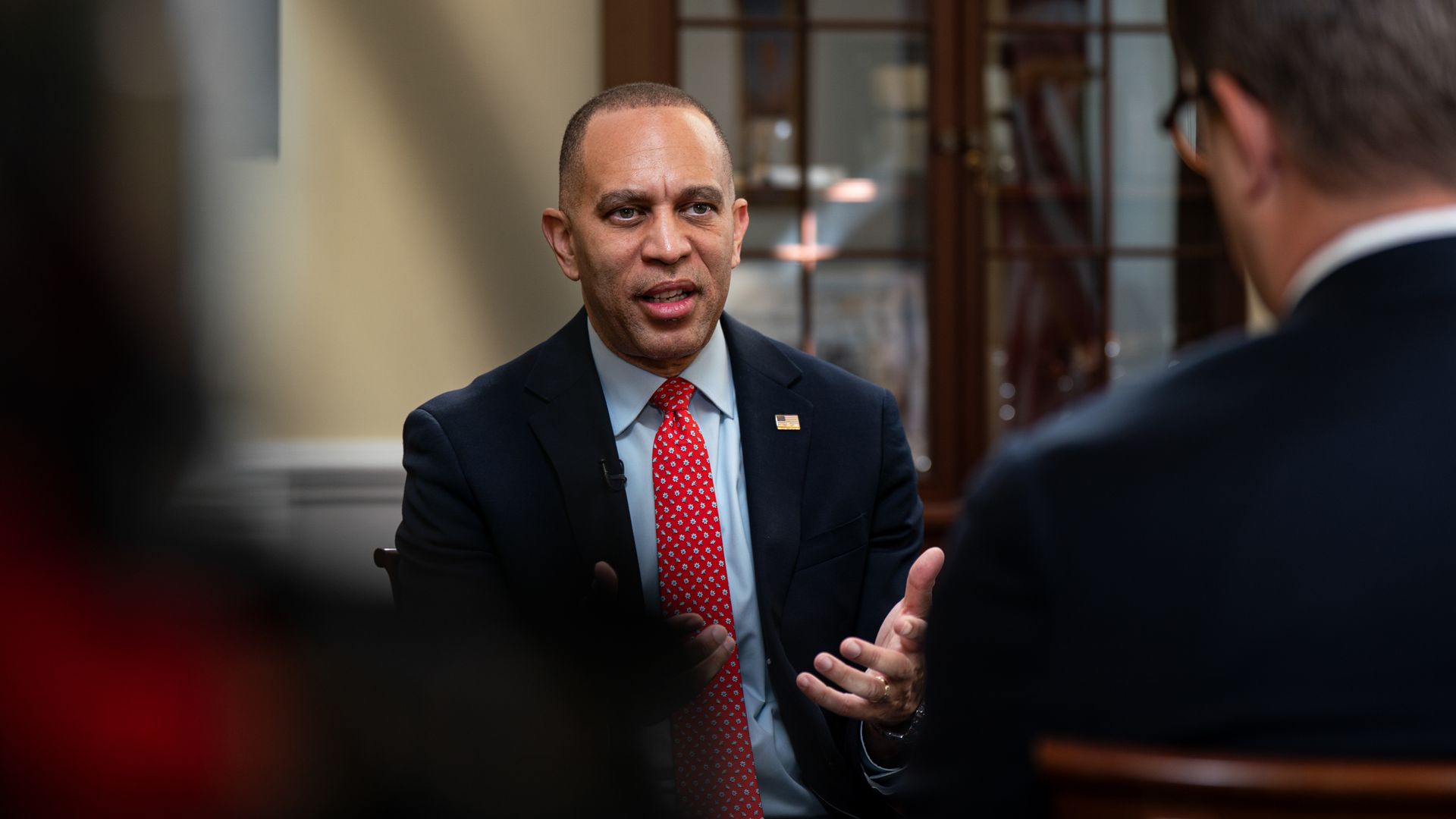 House Minority Leader Hakeem Jeffries, wearing a blue suit and a red tie, sits in a chair in front of a beige wall and a wooden cabinet.