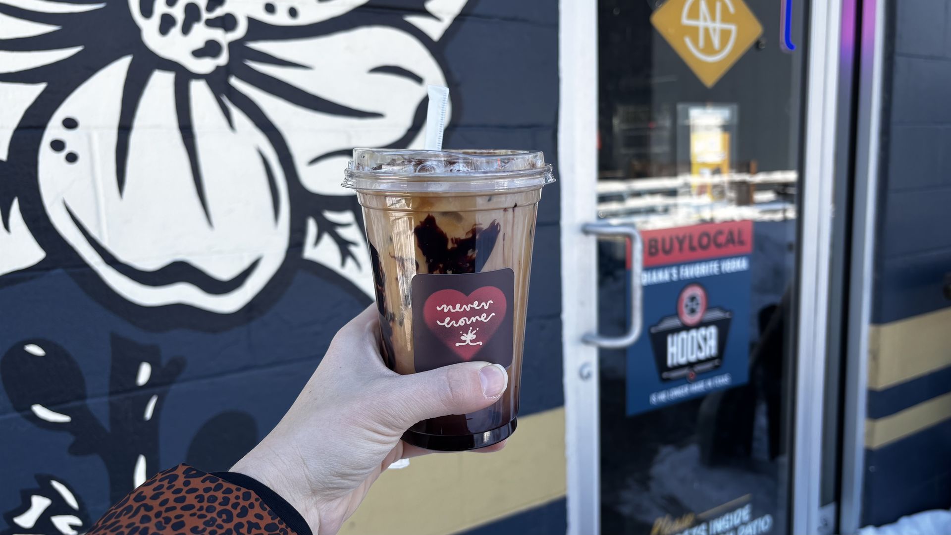 Hand holding iced coffee with chocolate drizzle and a "never home" heart logo cup in front of a building with black and white floral mural and a door with a buy local sign.