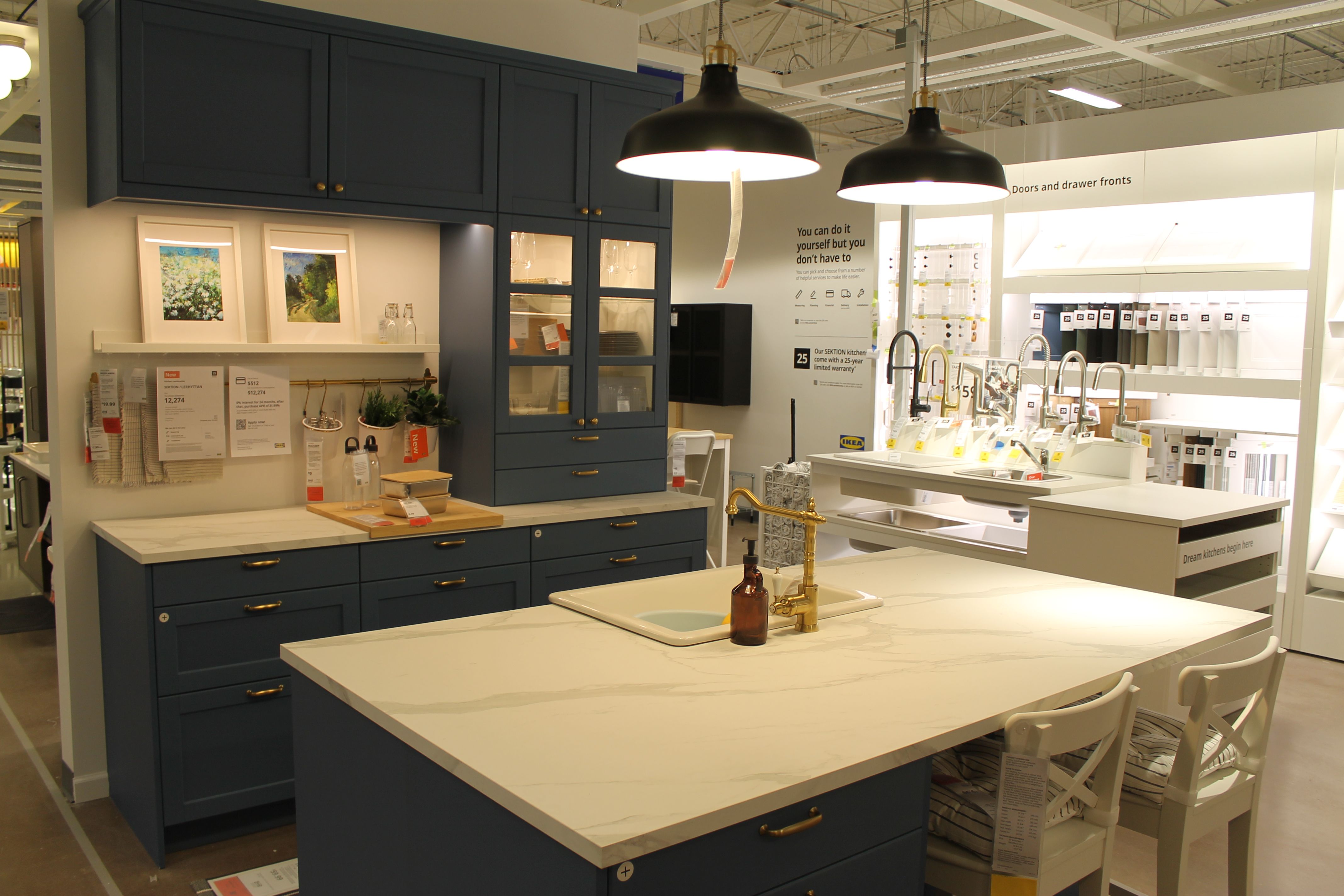 Modern kitchen showroom with navy blue cabinets, white marble countertops, gold faucet, two white chairs, two black pendant lights, and various kitchen displays and decor items.