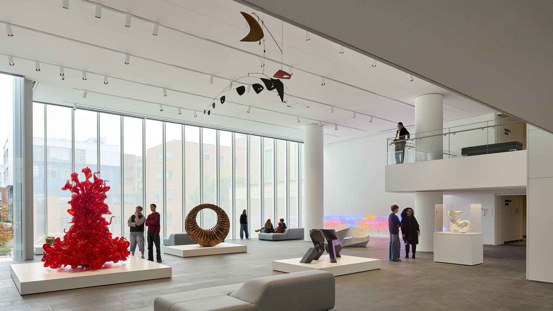 Modern art gallery with large windows showing city buildings. Red glass sculpture, circular brown sculpture, black abstract piece, white sculpture, neon text art, visitors viewing exhibits, and gray seating.