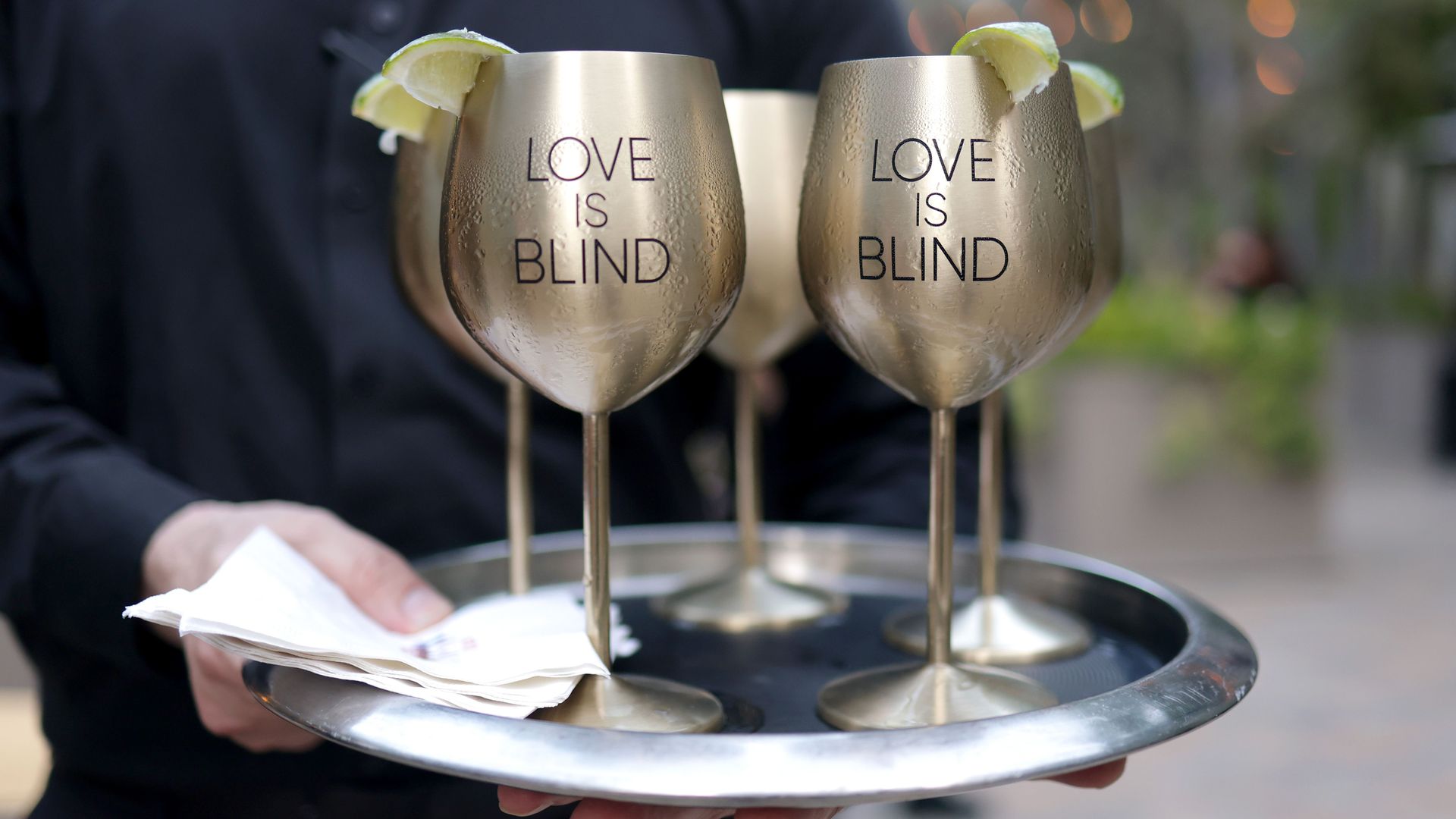 Four gold-colored wine glasses with lime slices are placed on a silver tray alongside napkins, with a hand holding the tray. Two of the glasses read LOVE IS BLIND." 