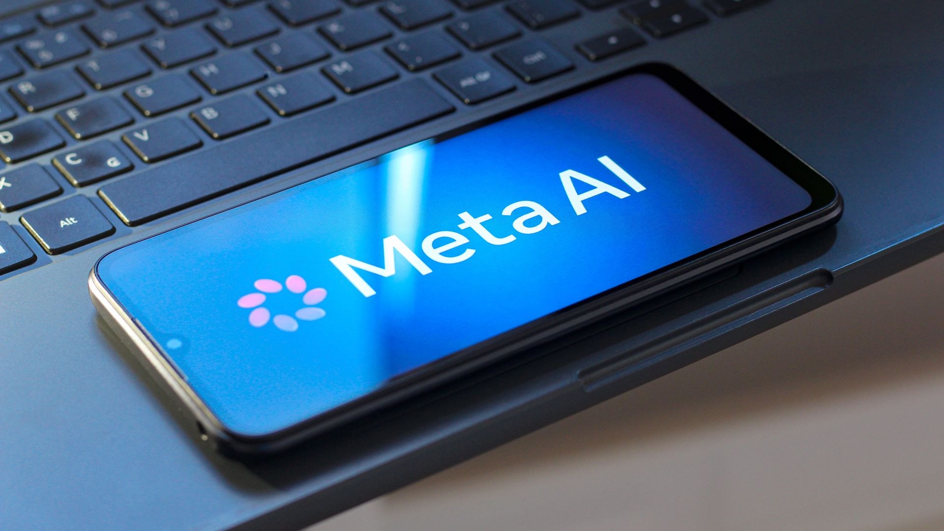 Close-up of a dark laptop keyboard with a smartphone resting on the lower edge; the phone screen shows a blue gradient with the white text Meta AI and a small circular logo.