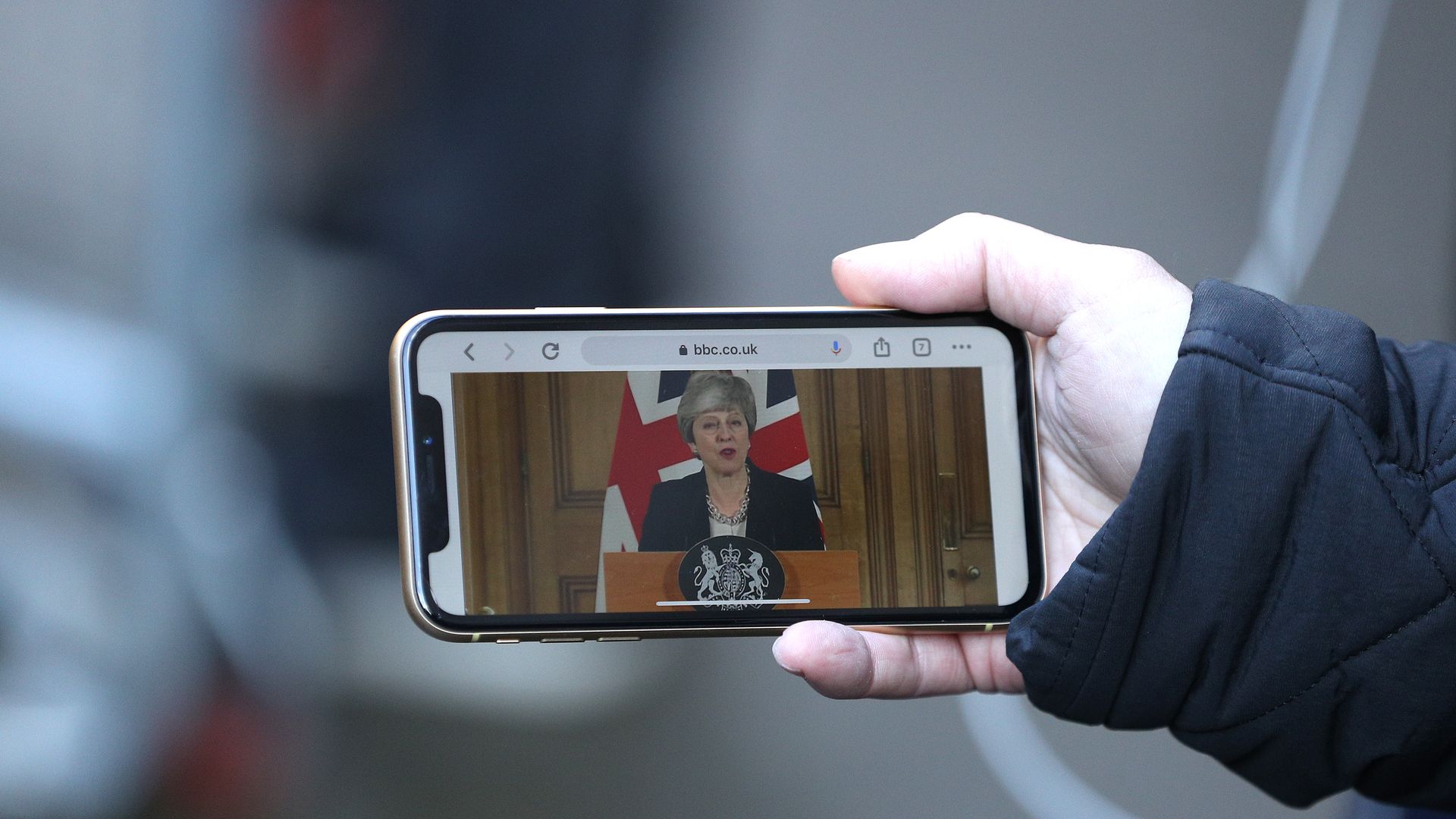 A man holds his phone, which is showing a video of British Prime Minister Theresa May.