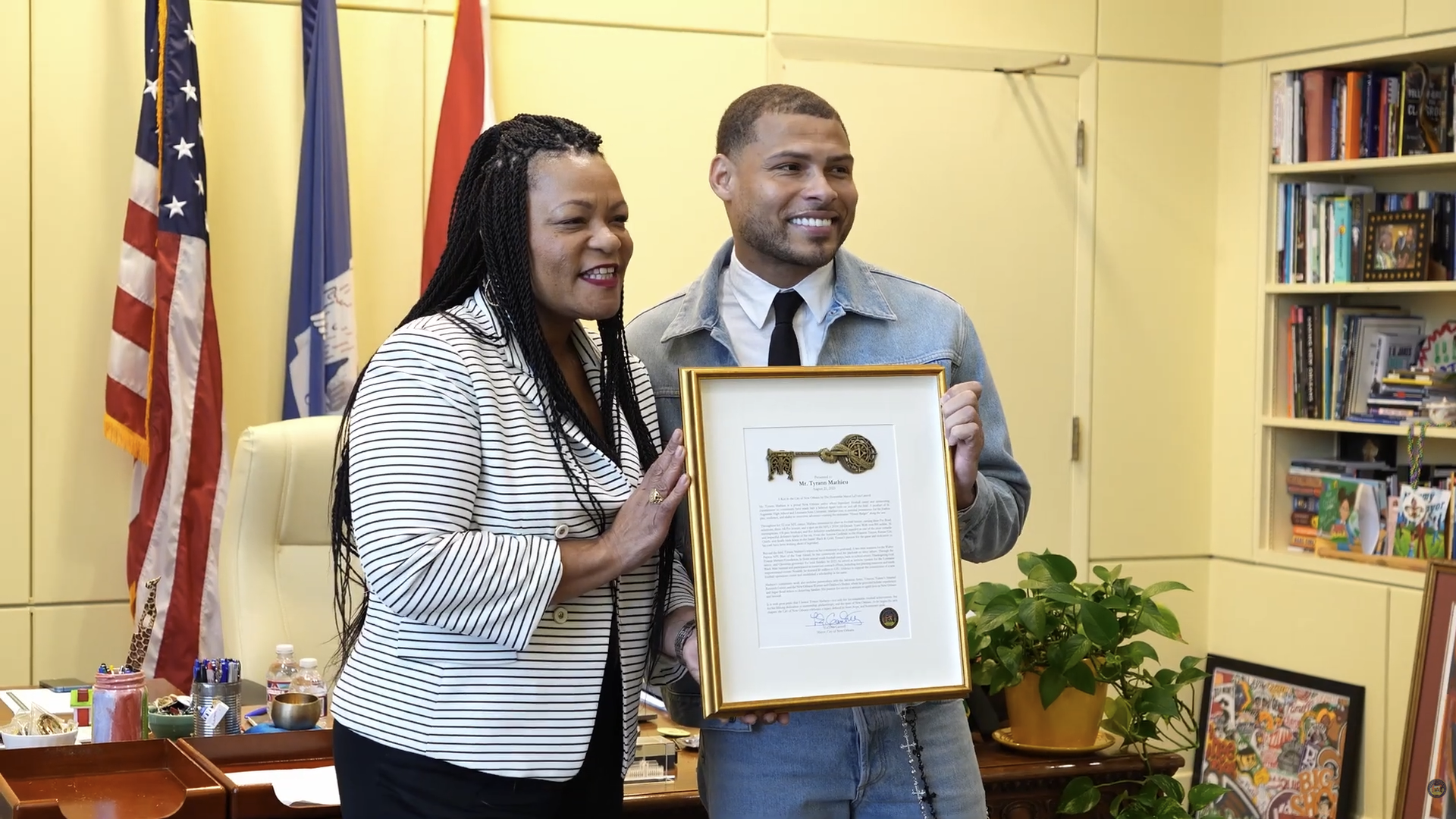 Two people smiling and holding a framed golden key award in an office with American and other flags, a desk, shelves with books, and a green potted plant.