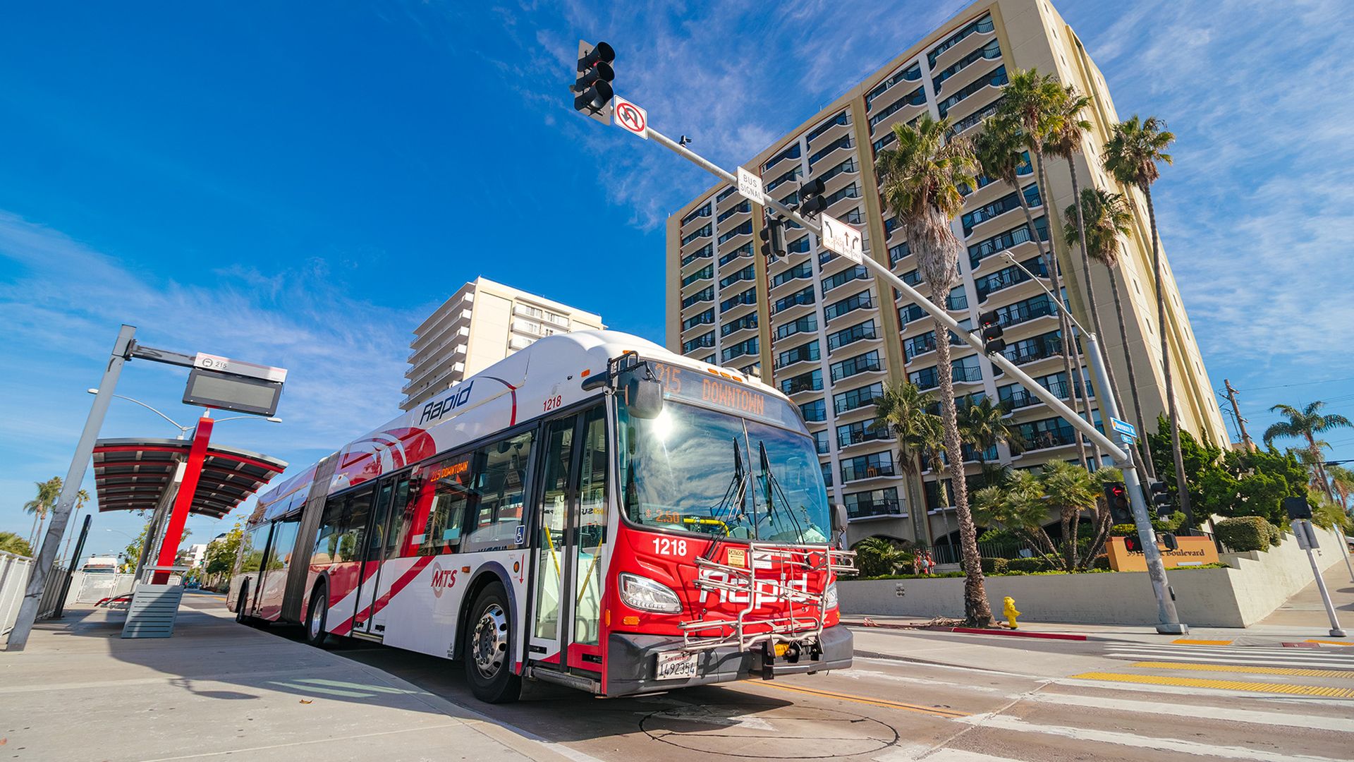 A red and white city bus labeled Downtown is parked near a tall beige high-rise, with palm trees and a bright blue sky behind. A diagonal traffic light pole and street signs hover above.