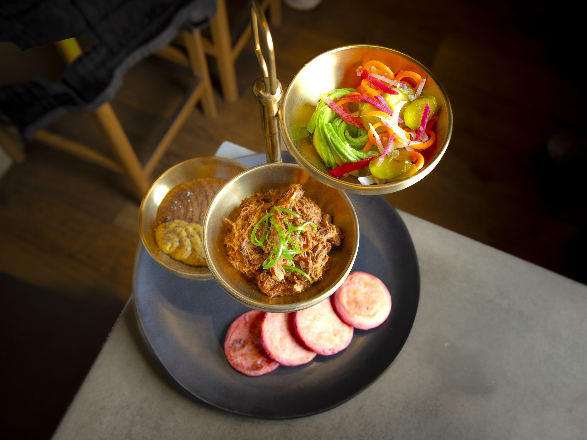 Three gold bowls with shredded meat, sliced avocado and pickled vegetables, and round crackers on a black plate with pink arepas on a table.