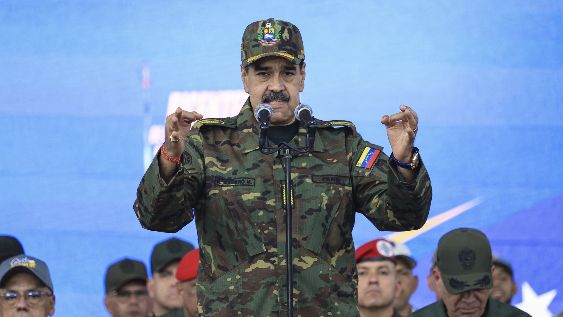 A man in camouflage military uniform and cap with a Venezuelan flag patch speaks at a podium with two microphones, gesturing with his hands, against a blue background. Several men in military attire stand behind him.