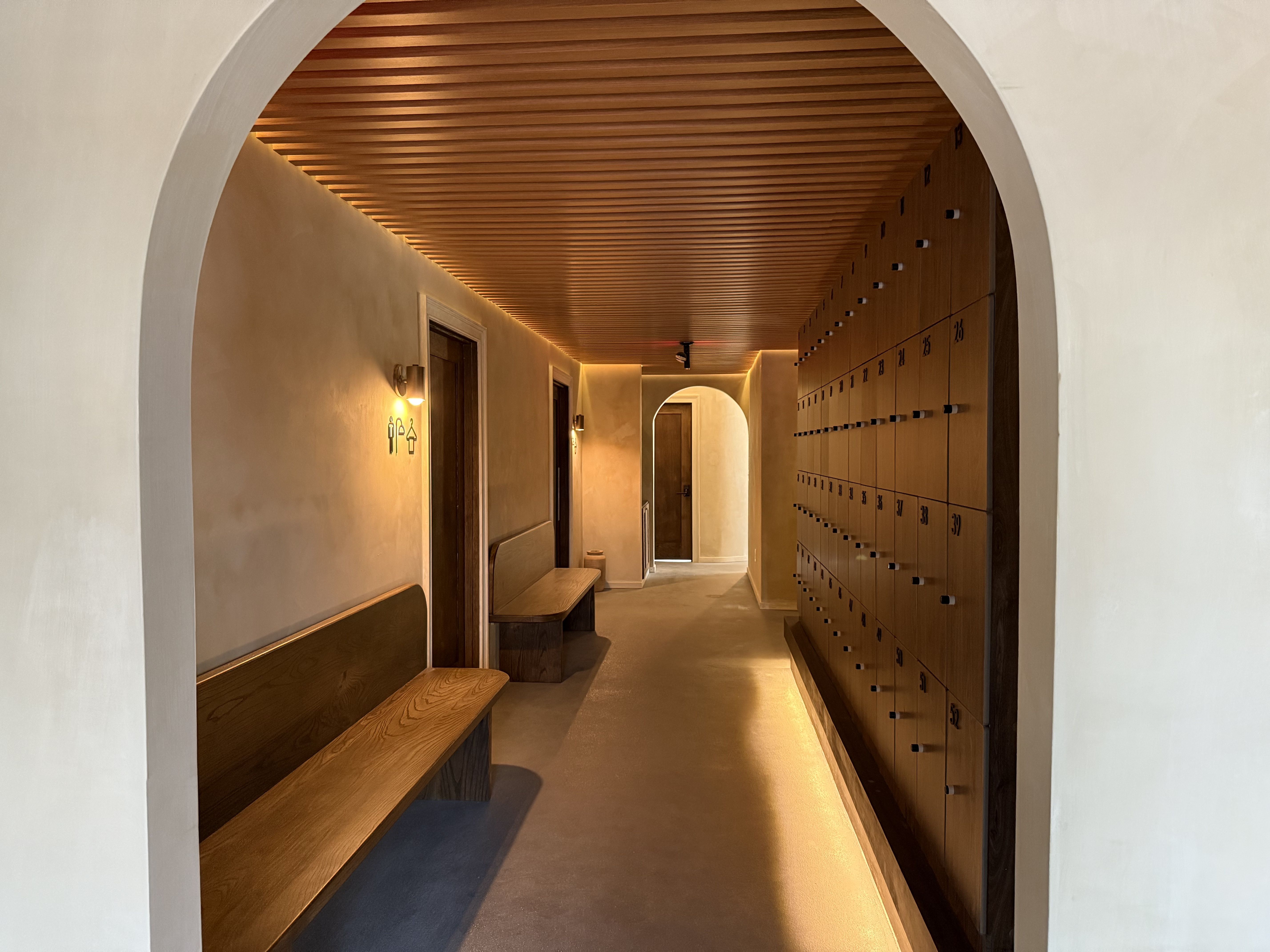 Hallway with wooden ceiling and benches on left, numbered wooden lockers with black handles on right, softly lit warm beige walls, and arched doorways.