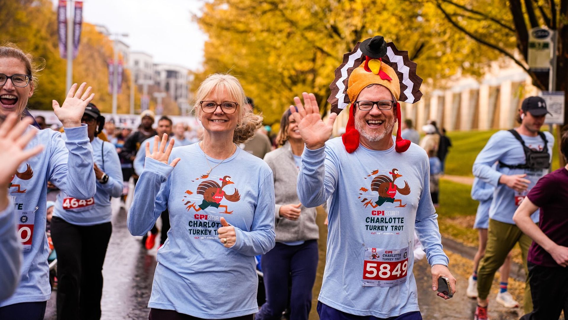 Smiling runners in light blue shirts with turkey logos wave while running in the Charlotte Turkey Trot race; one man wears a turkey hat; fall trees with yellow leaves line the street.