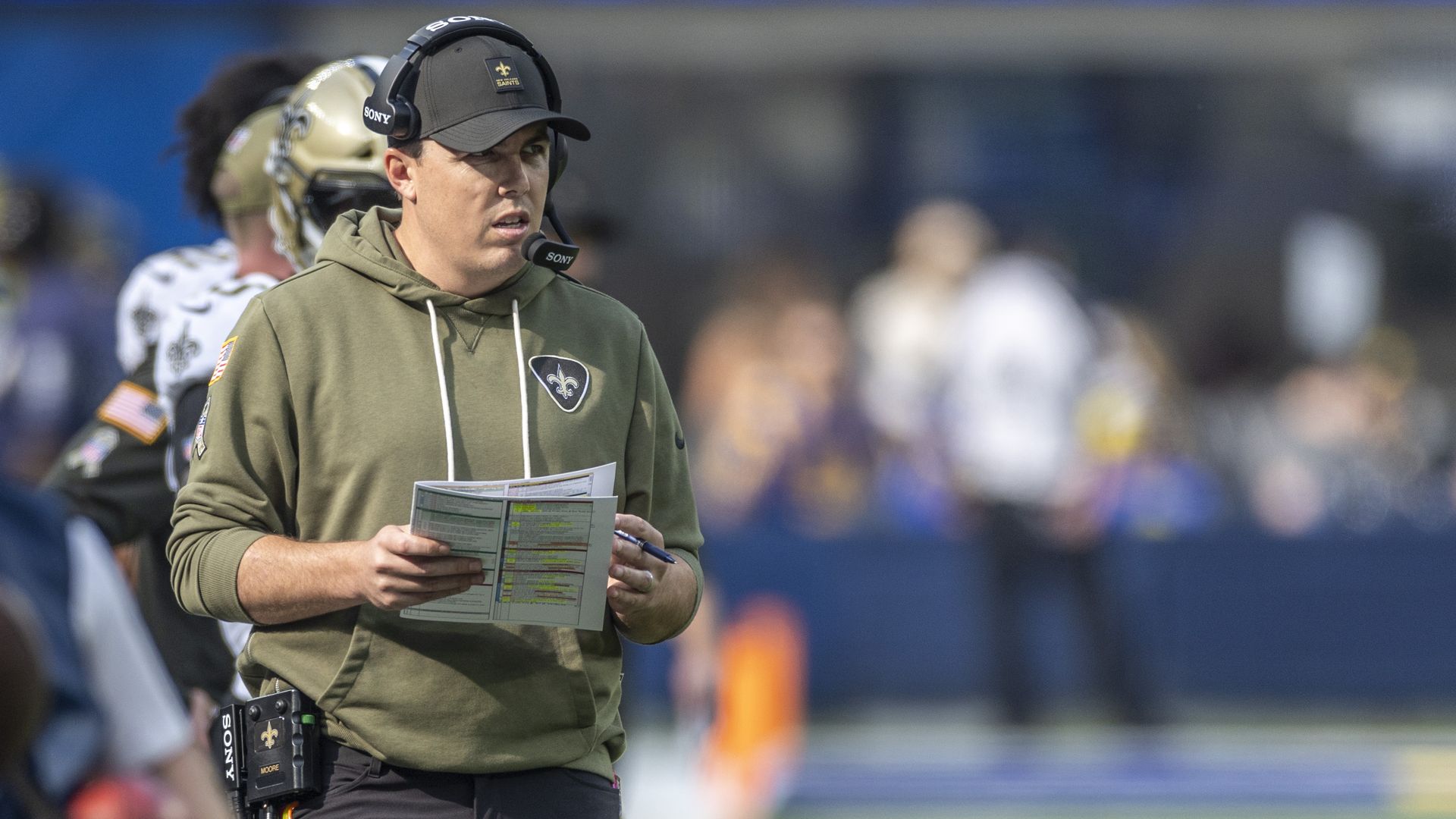 Kellen Moore, wearing a green Saints-branded hoodie and hat, holds a clipboard as he eyes a football field.