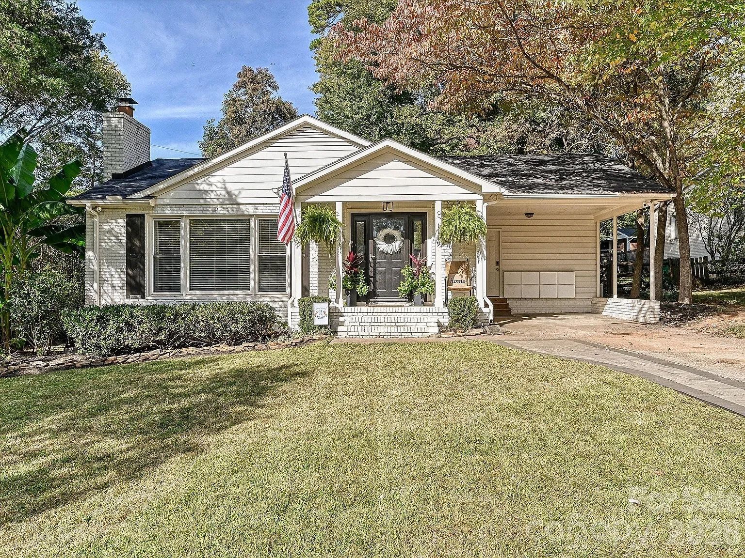White single-story house with black front door, American flag, hanging plants, and green lawn in front under a partly cloudy sky with trees around.