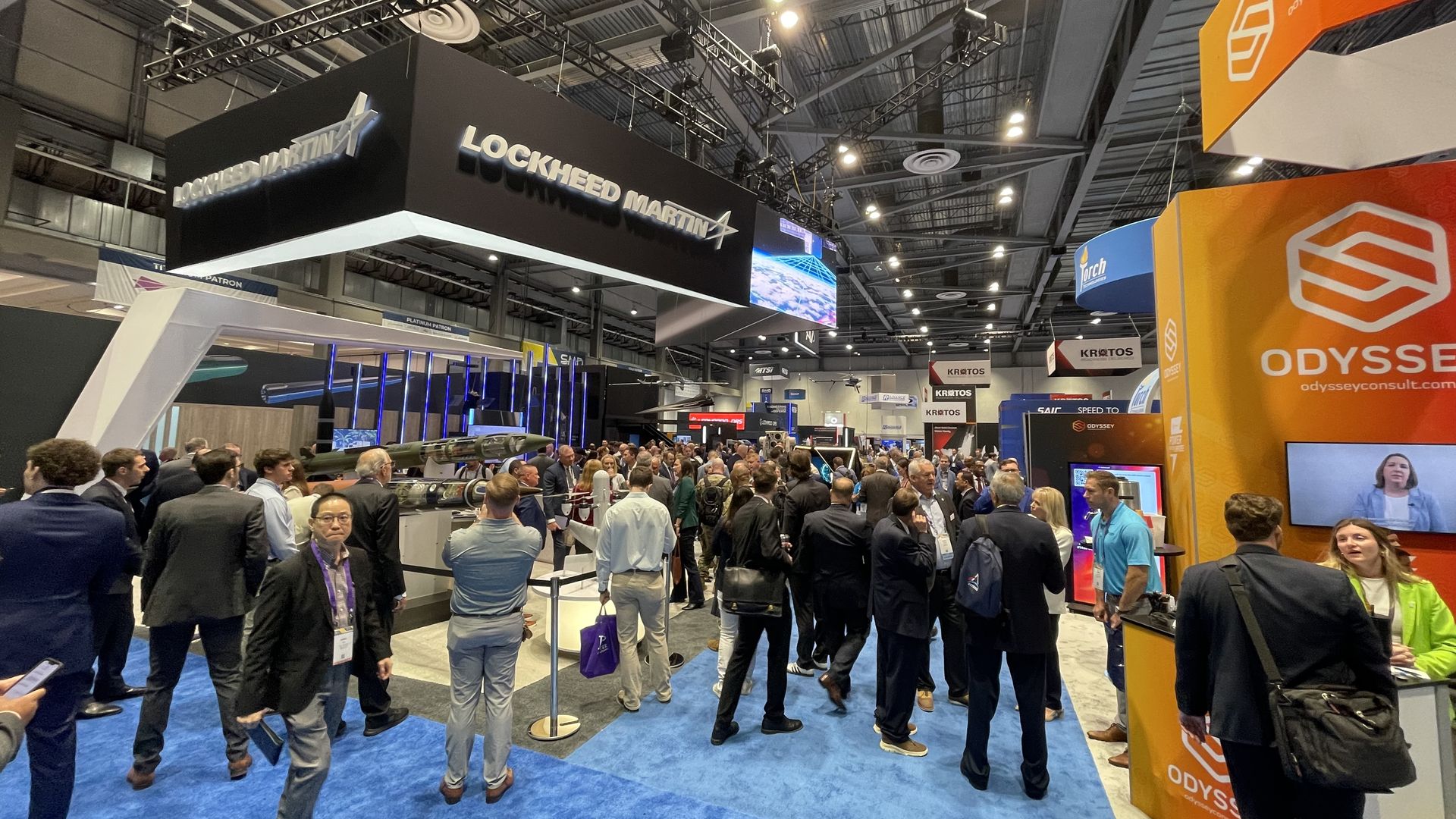 Busy trade show floor with a large black Lockheed Martin booth suspended from the ceiling, a dense crowd of professionals in suits, and a bright orange Odyssey display on the right; blue carpet.