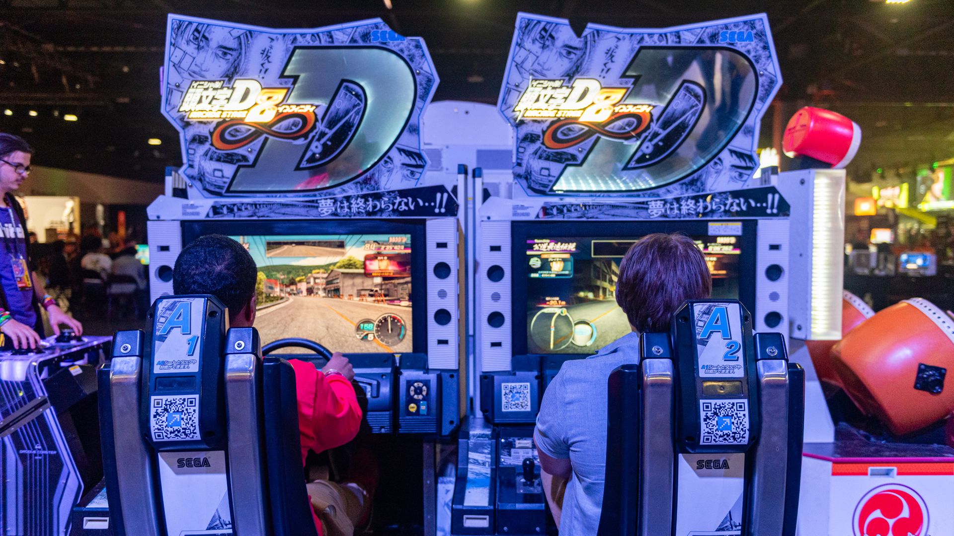 Two people playing side-by-side SEGA arcade racing games with blue and white game cabinets, in a dimly lit space with other games visible.