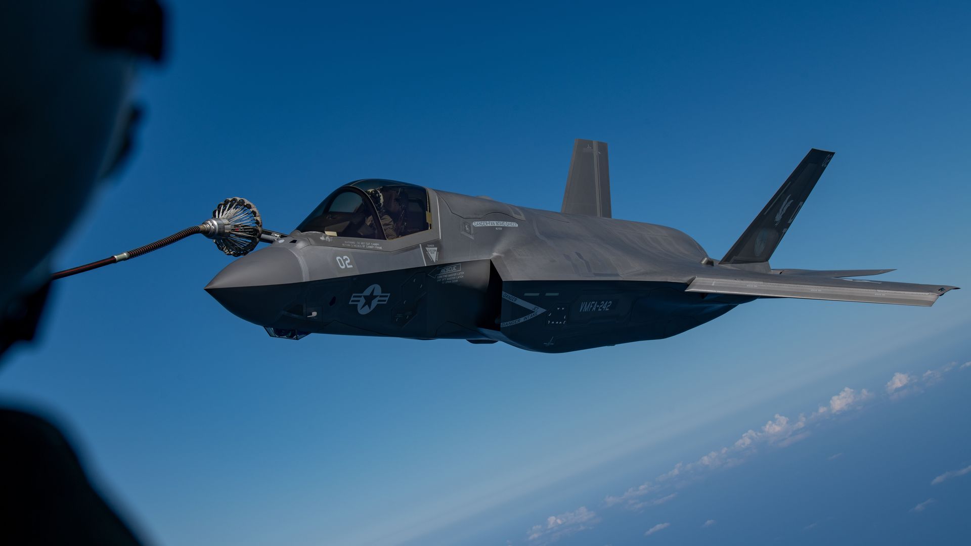 An F-35 lines up for aerial refueling. A person can be seen on the left edge of the frame. The sky is blue with few clouds.