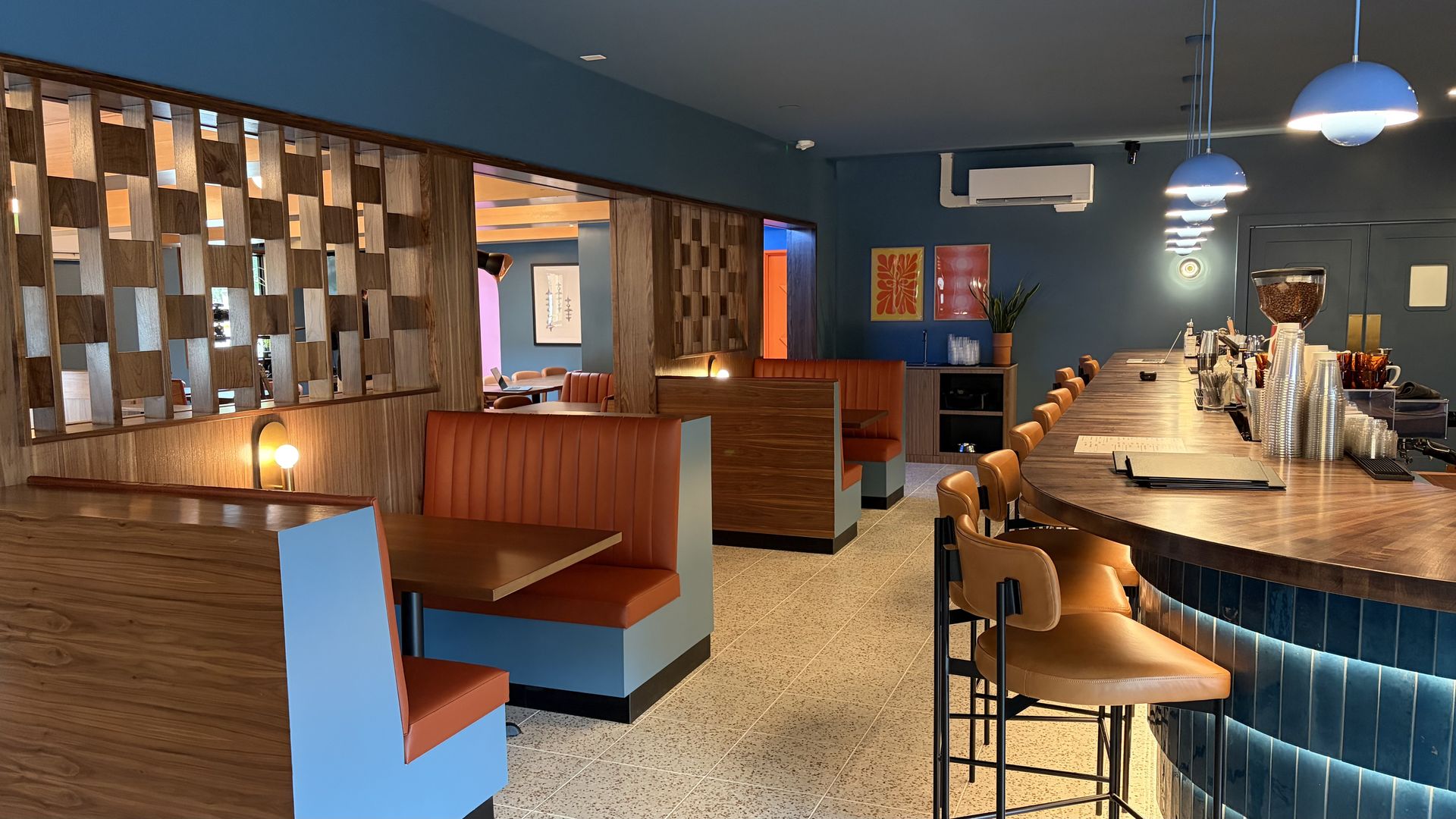 Interior view of a retro-modern cafe/bar with blue walls, orange booths, wooden lattice dividers, a long curved bar with stools, blue pendant lights, and a coffee station.