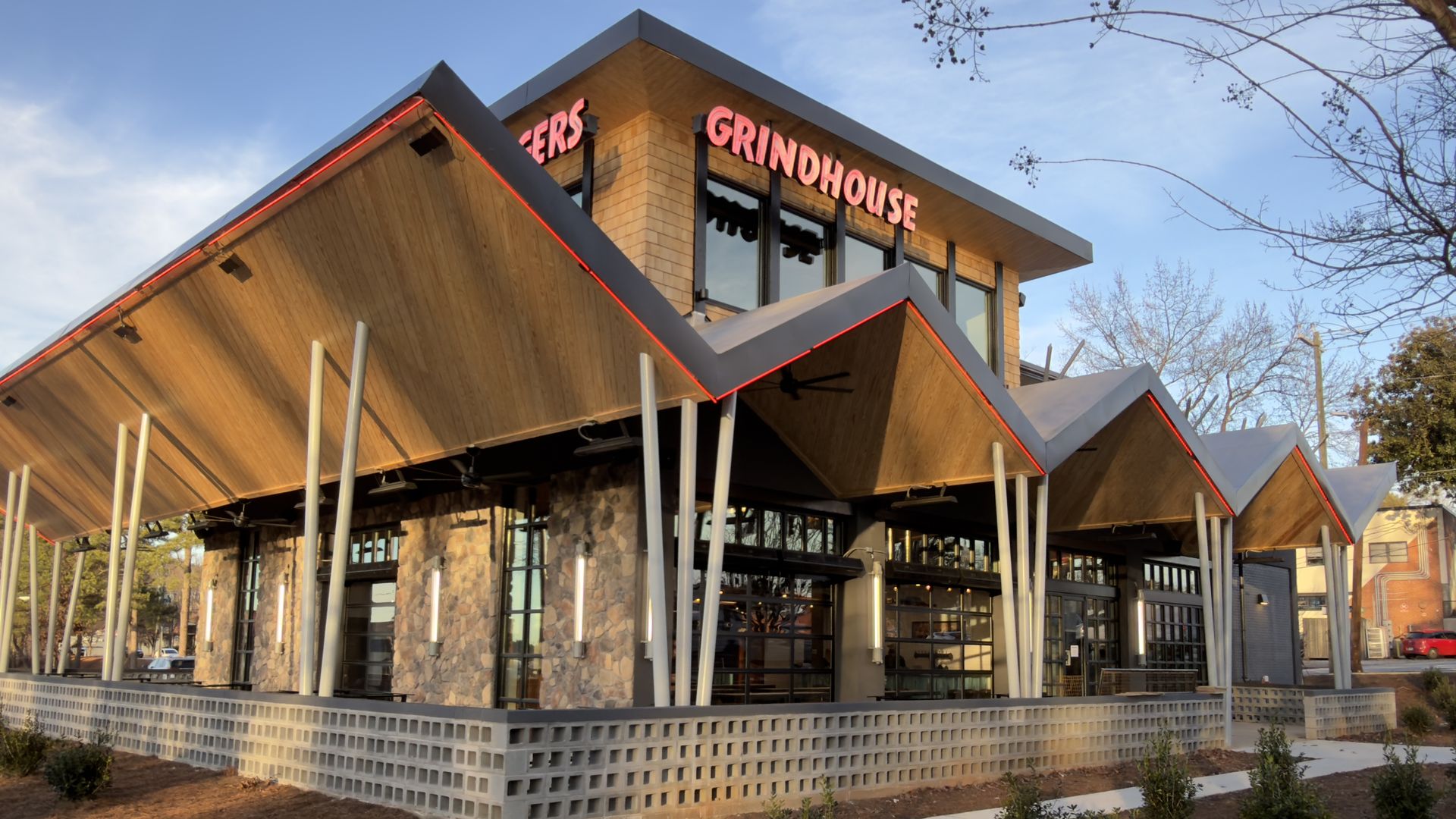 Modern building with a zigzag wooden roof and stone walls, neon red signs reading "GRINDHOUSE" and "BURGERS," under a clear blue sky with sparse trees around.