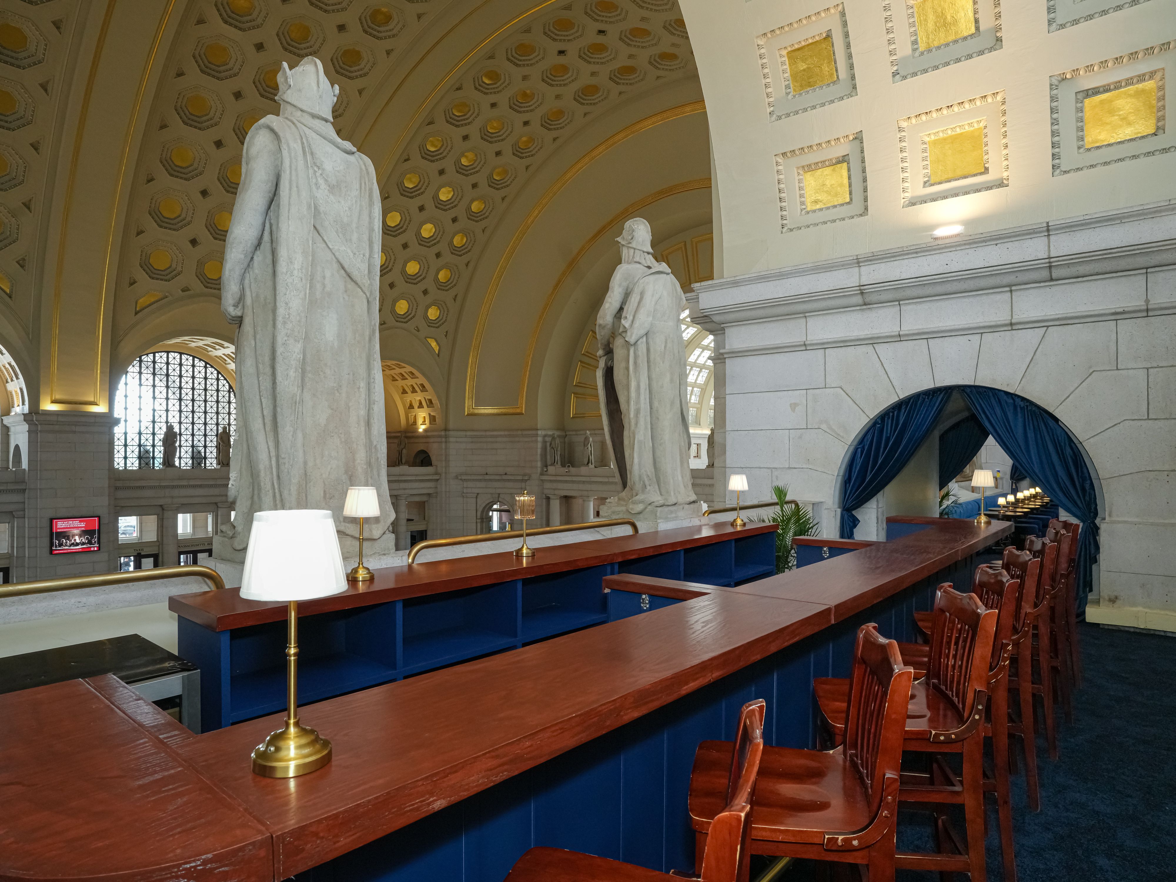 Interior of a grand hall with white marble statues facing away, ornate gold-accented coffered ceiling, tall arches, and a long wooden counter with brass lamps and blue base.