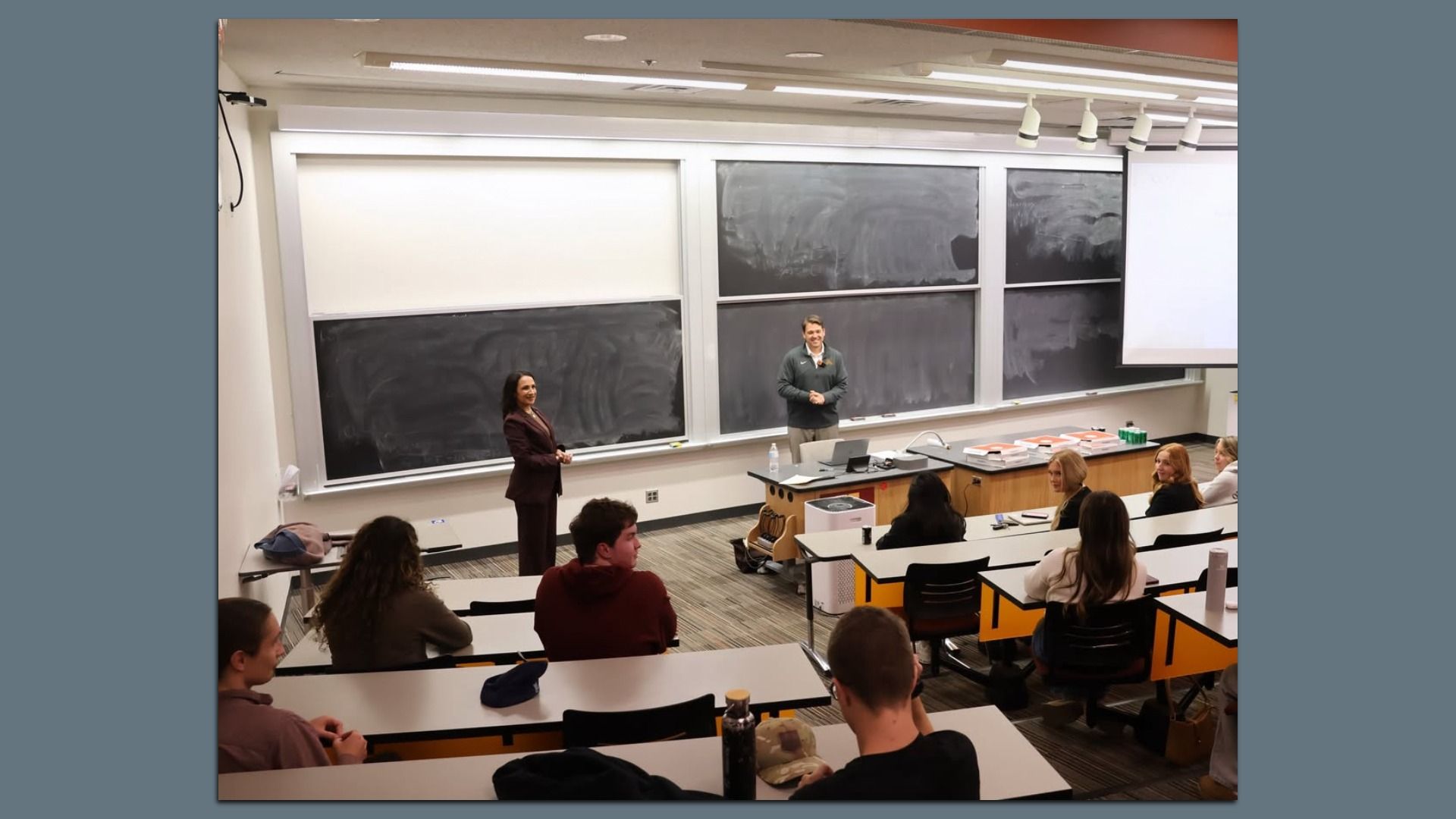 A university lecture hall with two presenters at the front: a woman in a burgundy suit by the left chalkboard and a man in a gray pullover at the center, while students sit at desks facing them.
