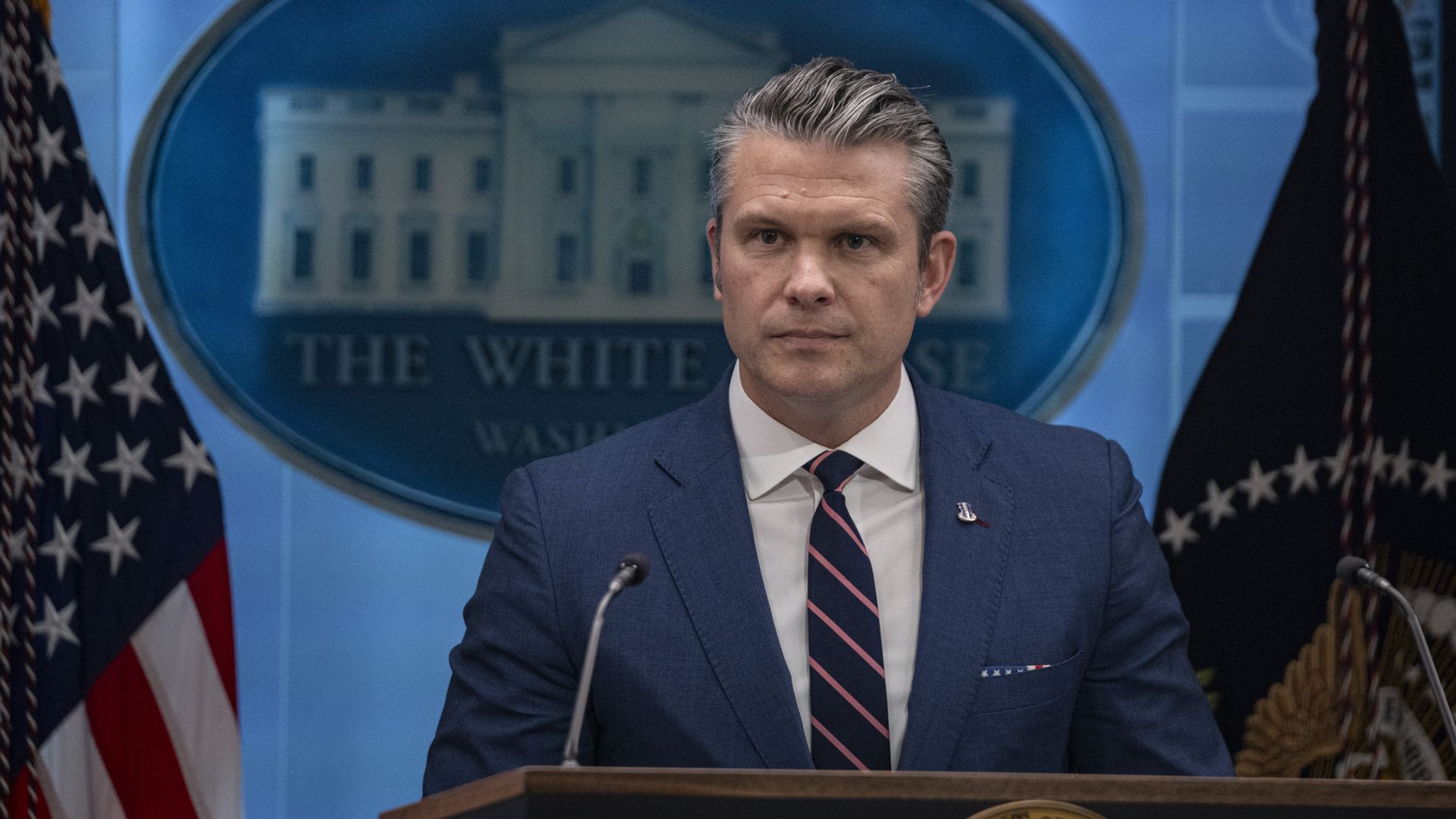 A man with gray hair in a navy suit and striped tie stands at a podium in the White House briefing room, with American flags and the White House seal visible in the background.