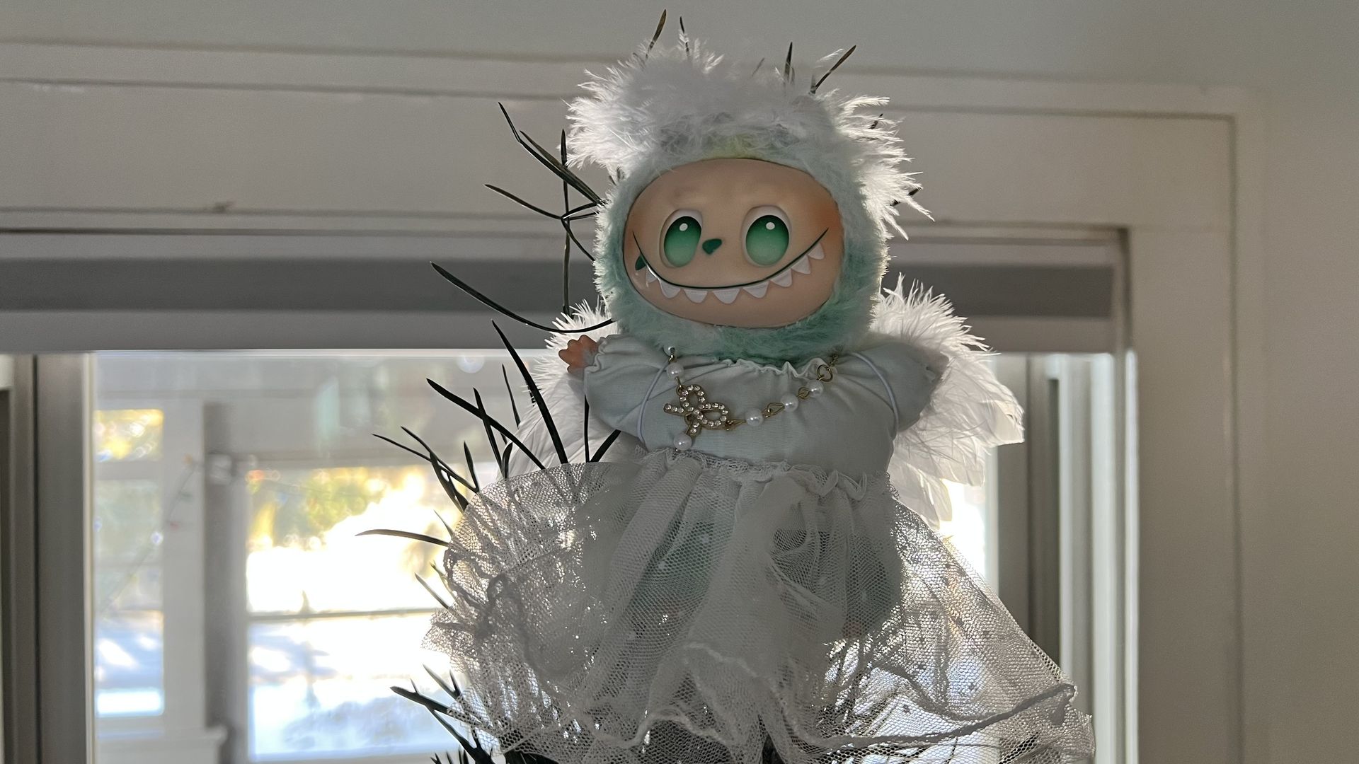 A labubu with big green eyes, a wide toothy grin, and a fluffy white feathered hood and dress, standing in front of a bright window with black twig-like decorations behind.