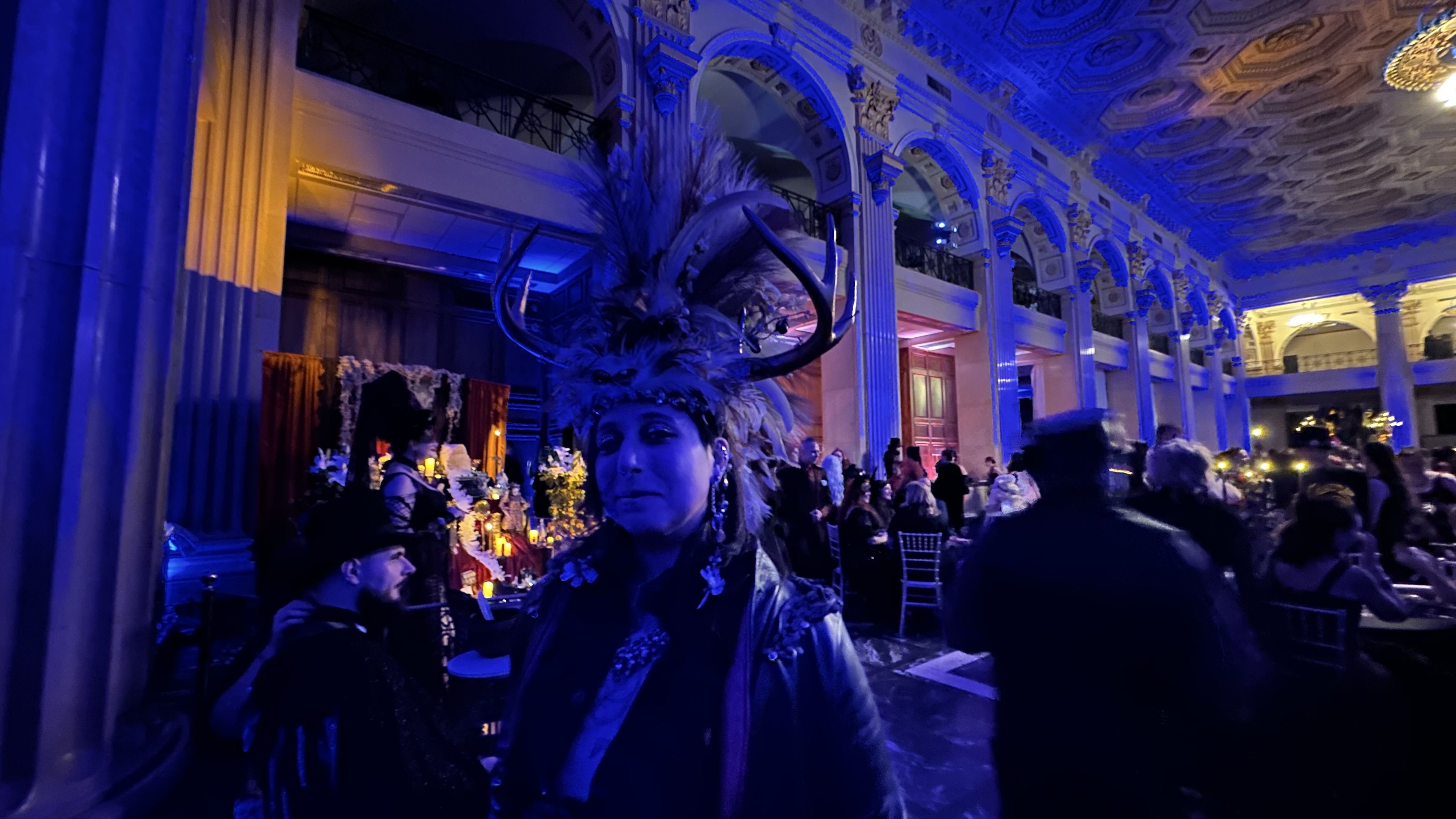 Indoor event in a grand hall with ornate arches and ceiling, blue lighting, people dressed in dark, elaborate costumes including a person wearing a feathered headdress with antlers.
