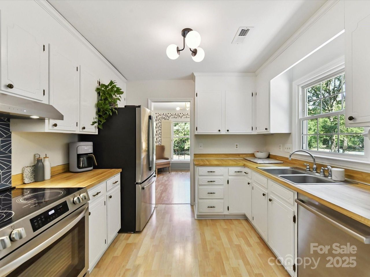 Bright kitchen with white cabinets, wood countertops, stainless steel appliances, a plant on the fridge, and a window showing green trees outside.