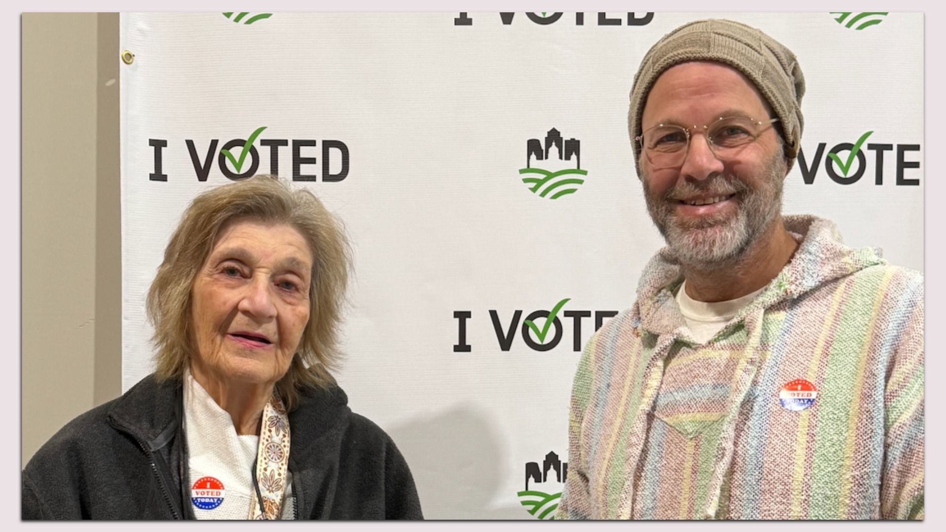 Two people pose in front of a white backdrop that reads "I Voted" with green check marks; an elderly woman in a dark coat on the left and a smiling man wearing a beige knit hat and glasses on the right.