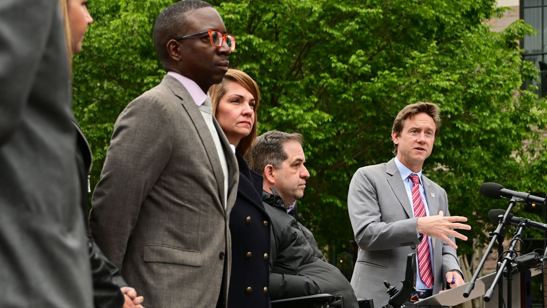A man speaks at a lectern while three other people stand nearby. 