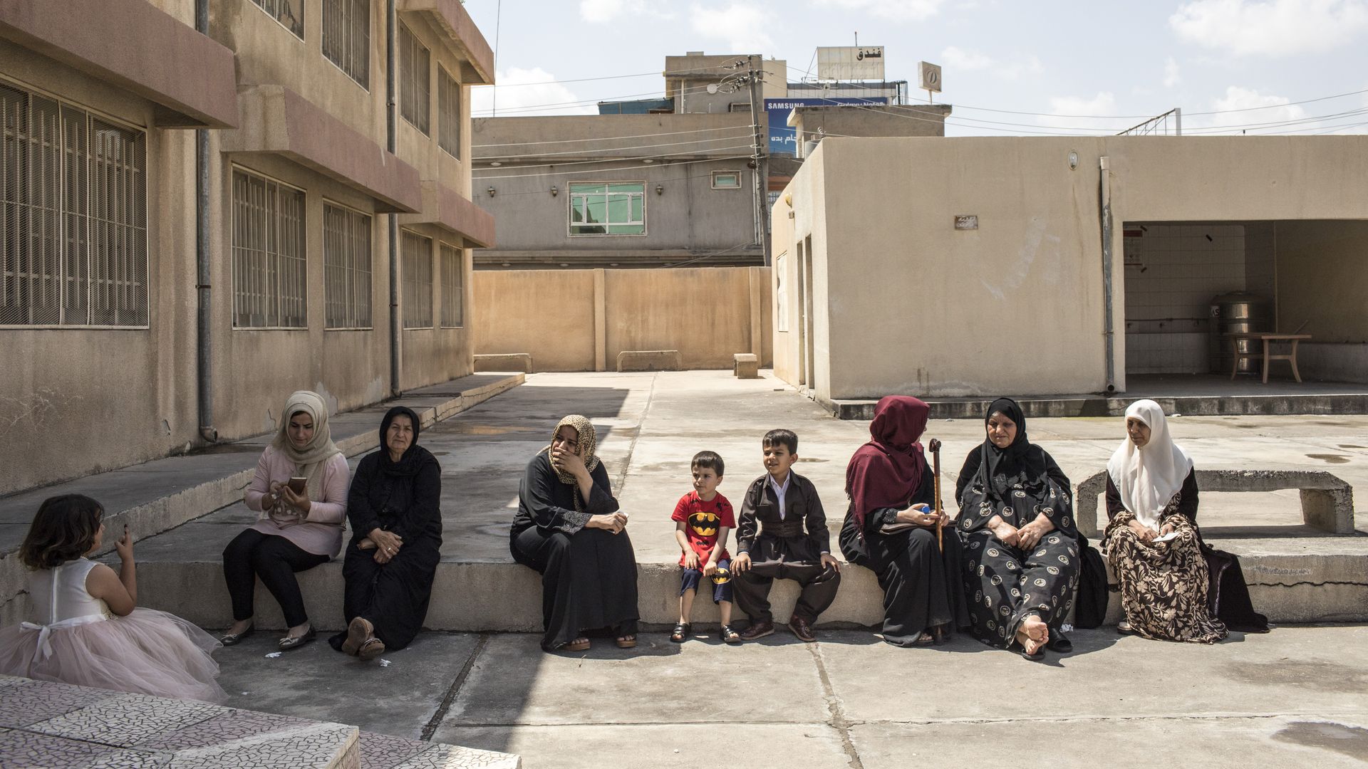 Women and children sit on a curb next to a building