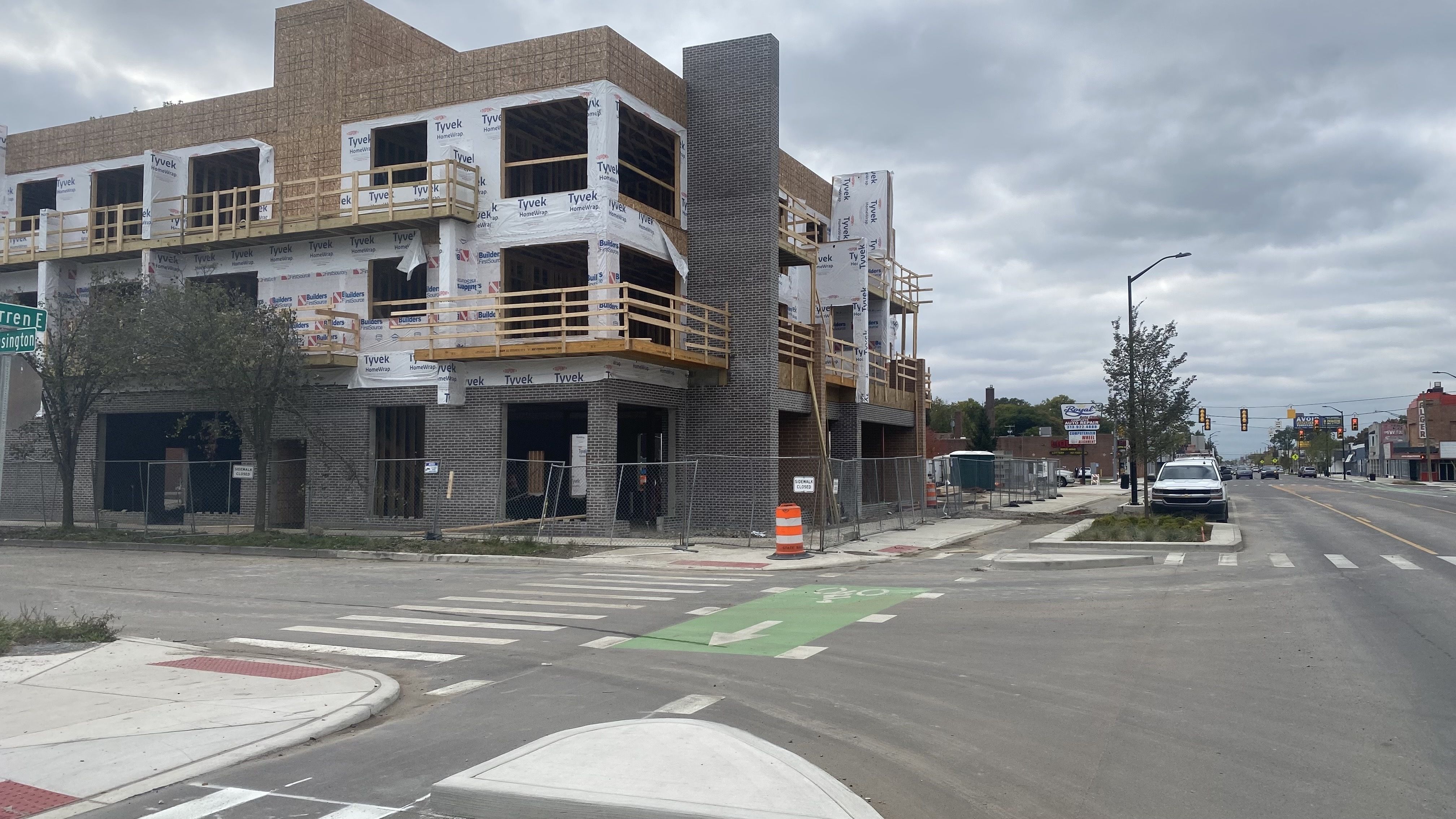 A three story building that's still under construction, alongside a street with new walkways and a new protected bike lane.