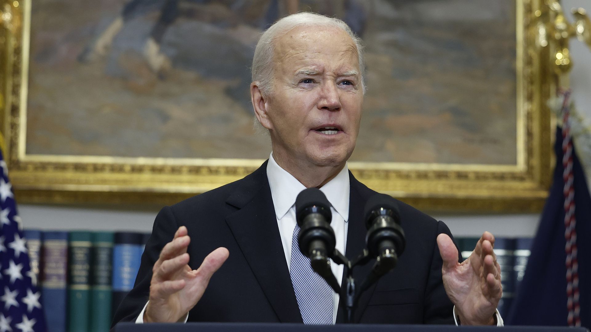 President Biden stands at a podium with his hands in motion and a framed painting and books behind him.